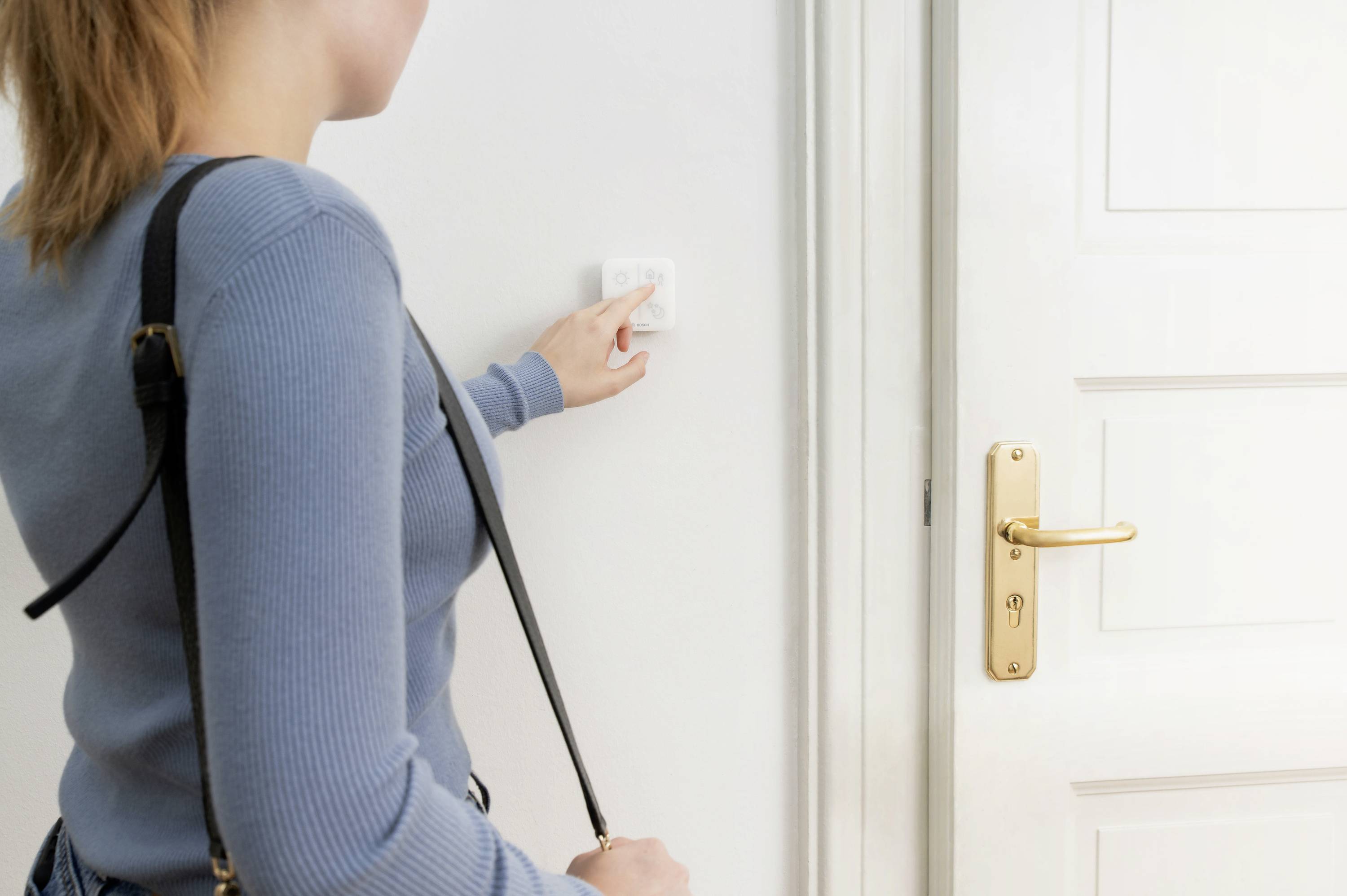 Person drückt einen Lichtschalter neben einer weißen Tür. Sie trägt eine blaue Strickjacke und eine schwarze Umhängetasche.