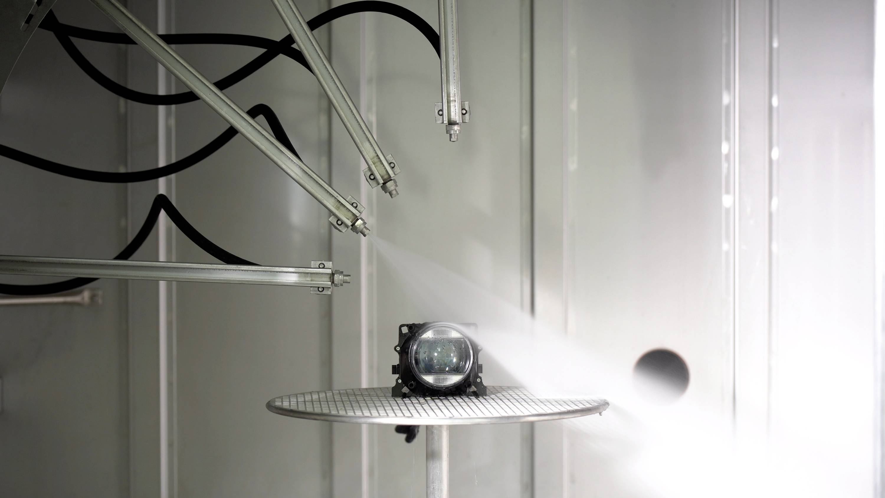 Ein kleiner Projektor auf einer runden Plattform wird von mehreren Düsen aus unterschiedlichen Winkeln mit Wasser besprüht.