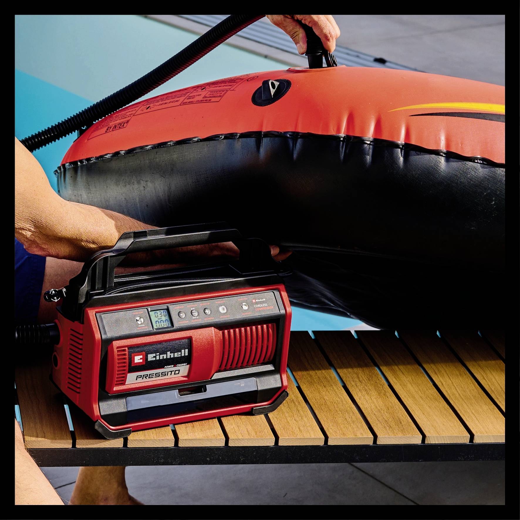 Eine Person pumpt einen großen roten Schwimmreifen mit einem tragbaren Luftkompressor auf. Der Kompressor steht auf einem Holztisch.