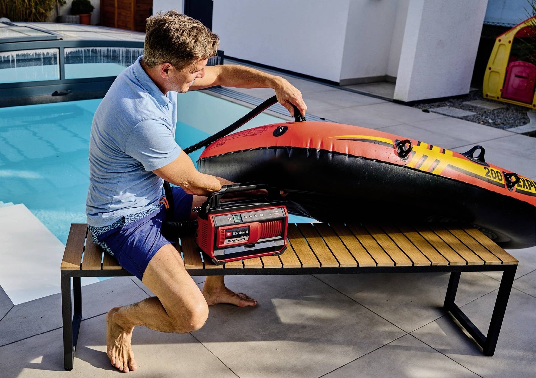 Ein Mann pumpt ein großes, orangefarbenes Schlauchboot mit einer tragbaren Luftpumpe auf, während er auf einer Bank neben einem Pool sitzt.