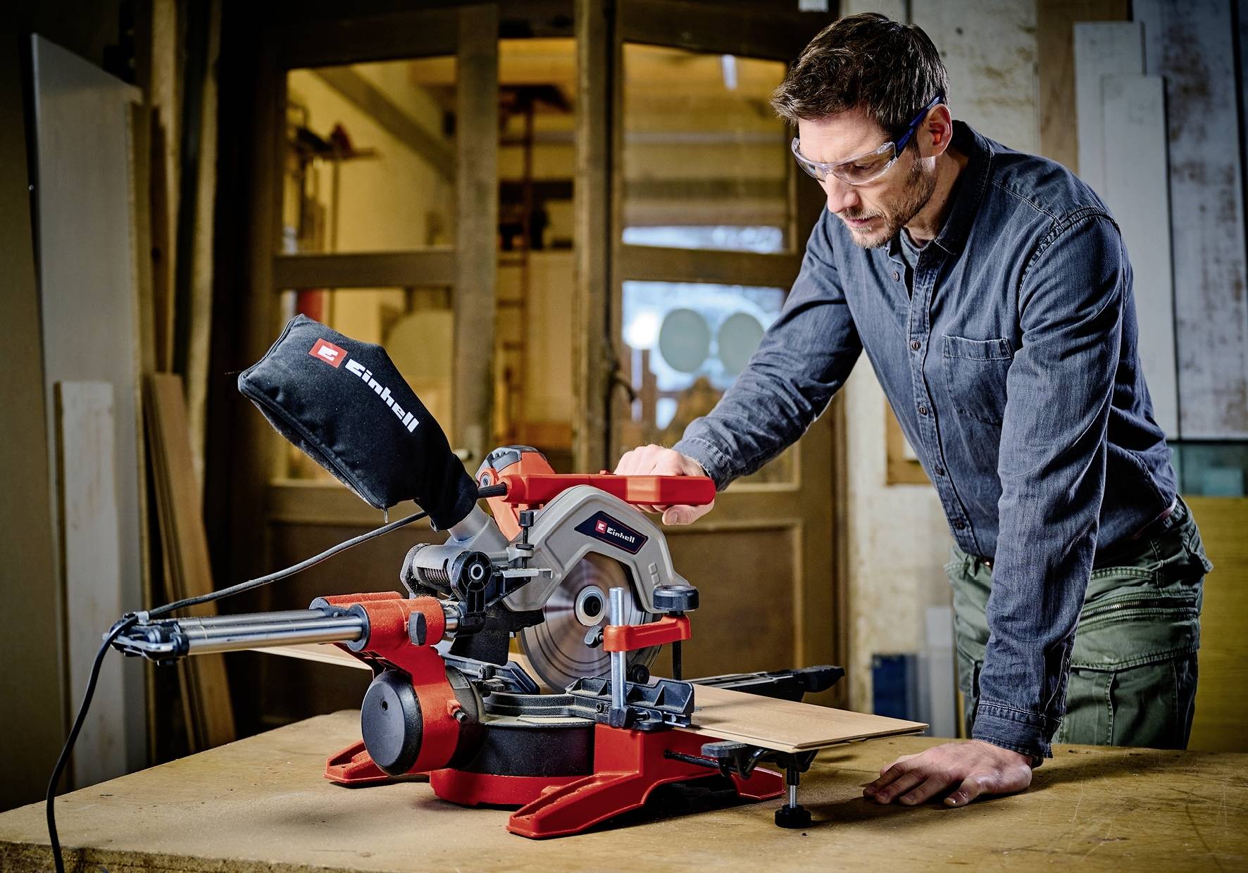 Ein Mann arbeitet mit einer Kappsäge, gekleidet in Schutzbrille und Hemd. Die Werkstatt im Hintergrund zeigt Holzbretter und Werkzeuge.