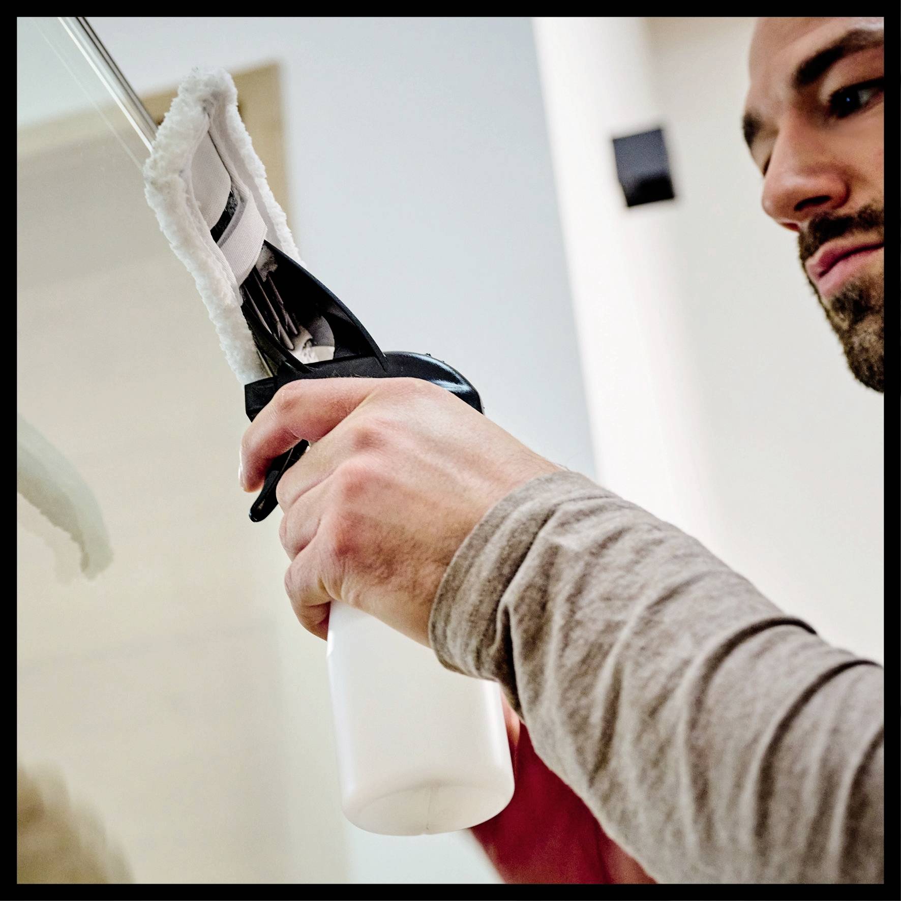 Mann reinigt ein Fenster mit einem weißen Spray und Tuch. Nahaufnahme auf die Hände. Sauberkeitskonzept in einem modernen Raum.