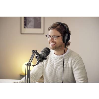 Person mit Kopfhörern und Mikrofon, sitzt lächelnd an einem Tisch, beteiligt sich an einer Audioaufnahme oder einem Podcast in einem gemütlichen Raum.