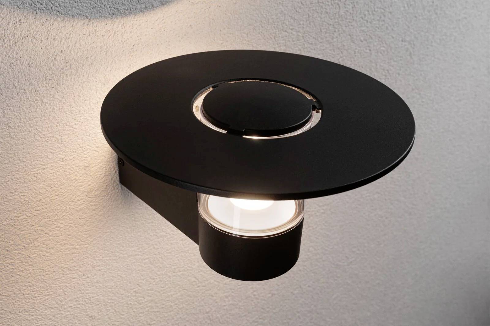 Schwarze, moderne Wandlampe mit rundem Schirm, leuchtet gegen graue Wand.