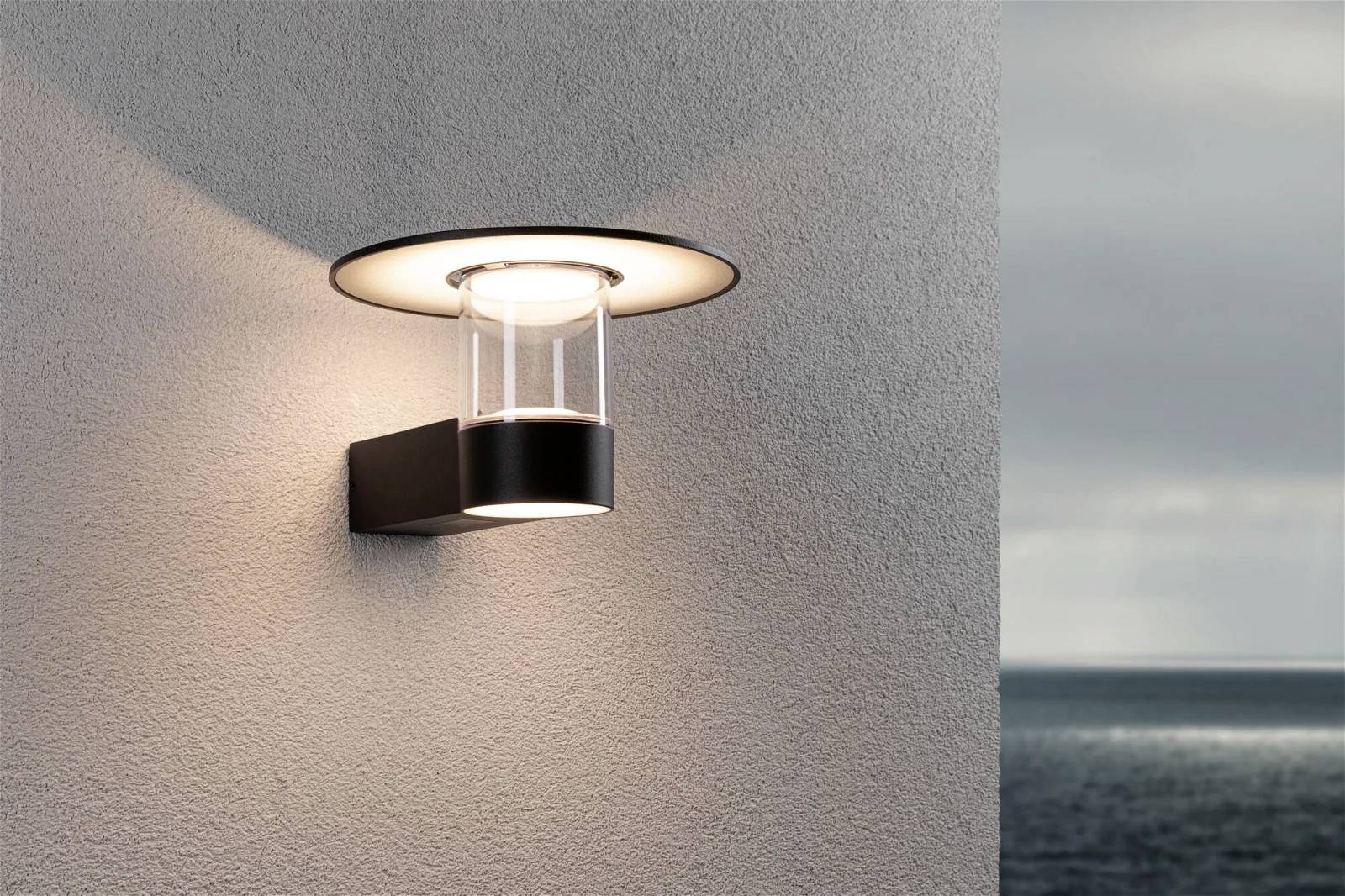 Moderne Wandaußenleuchte bei Dämmerung an einer Wand installiert, beleuchtet die Umgebung, mit Blick auf ein ruhiges Meer.