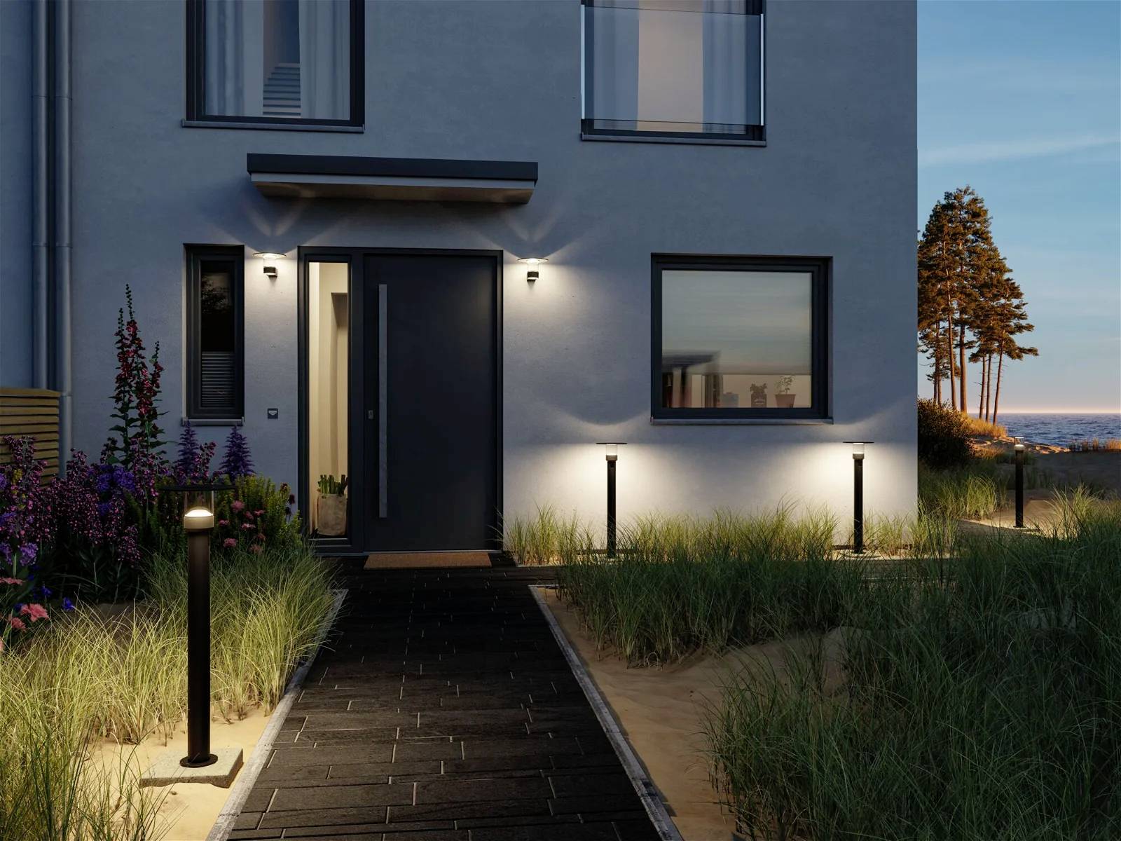 Ein modernes Haus bei Abenddämmerung, beleuchtet durch Außenlampen. Ein Weg führt zur Eingangstür, umgeben von Büschen und Gräsern.