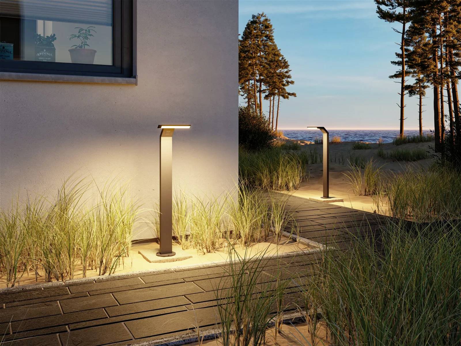 Gartenweg bei Sonnenuntergang, gesäumt von leuchtenden, modernen Wegleuchten. Links das Haus, im Hintergrund Bäume und Meer.
