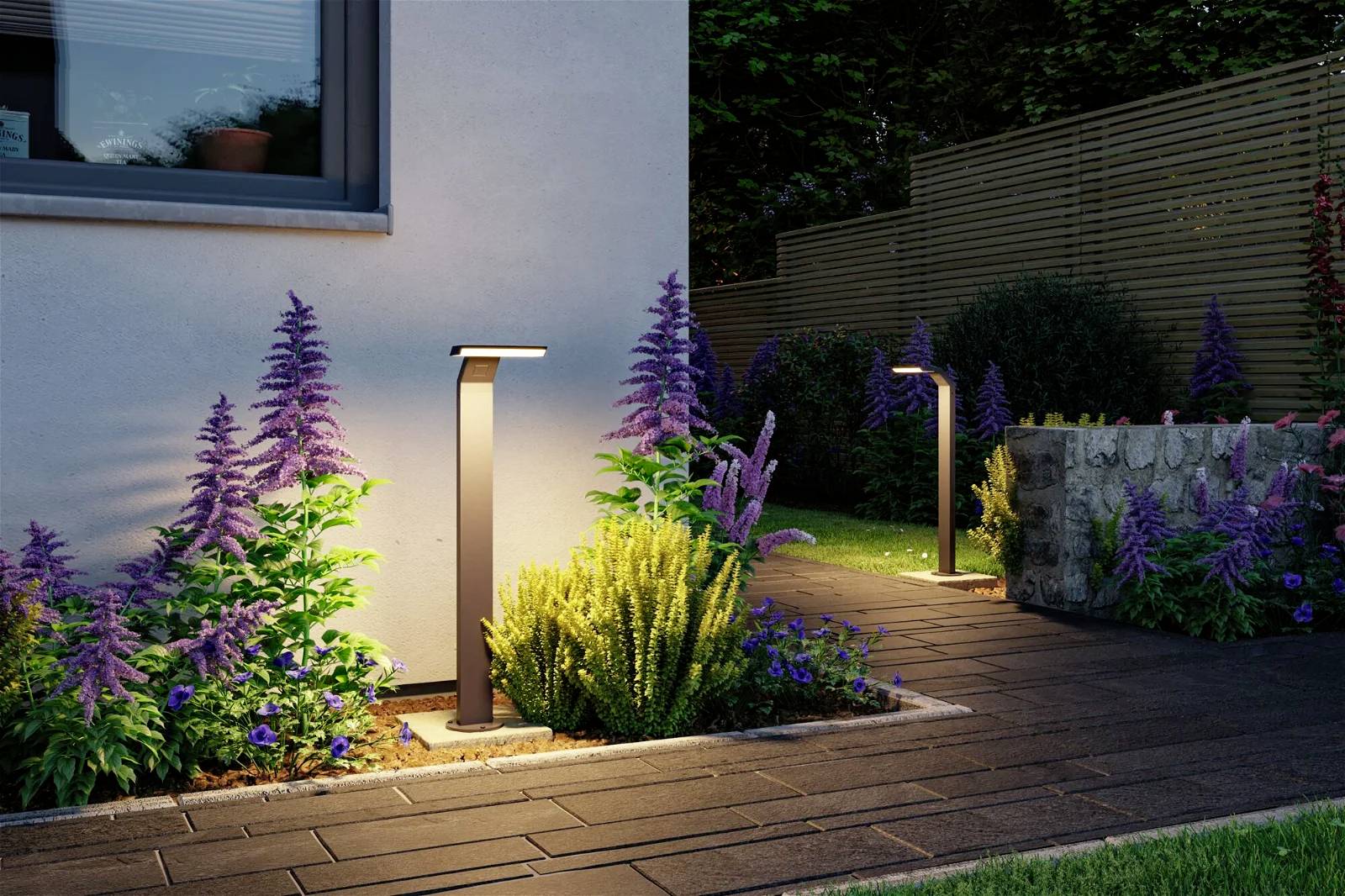 Beleuchteter Gartenweg bei Dämmerung mit modernen Wegleuchten und üppigen lila-blühenden Pflanzen entlang eines gepflasterten Pfades.