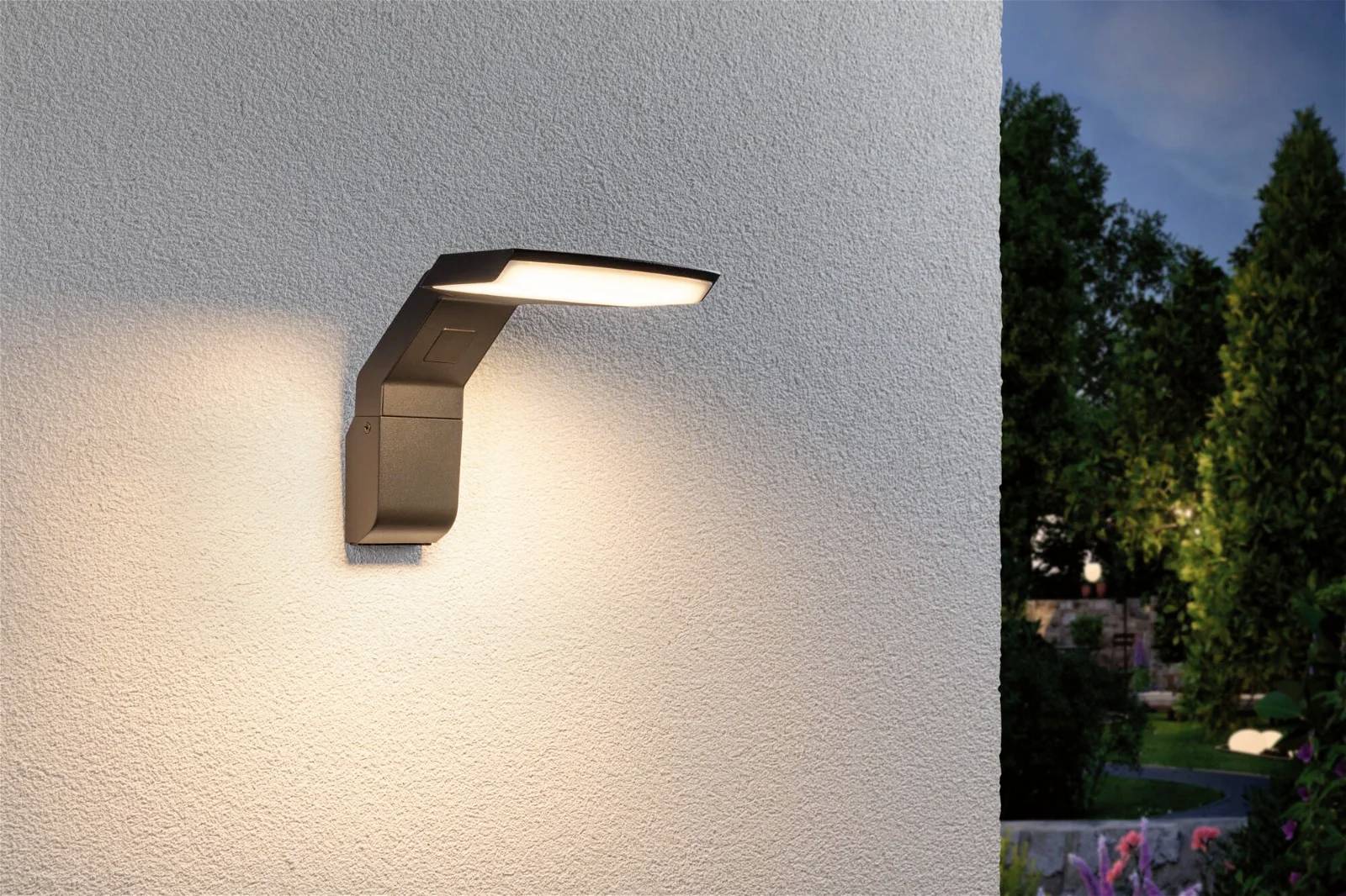 Moderne Außenwandleuchte an einer Hauswand, die warmes Licht abstrahlt. Im Hintergrund sind Bäume und ein Teil eines Gartens sichtbar.