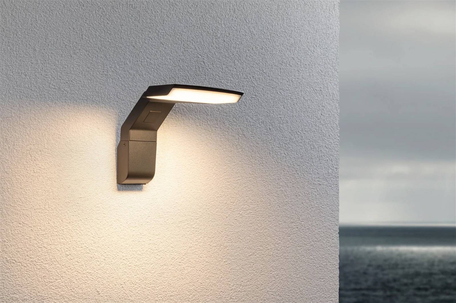 Wandleuchte an einer weißen Mauer, beleuchtet die Umgebung. Im Hintergrund ist das Meer unter einem bewölkten Himmel sichtbar.