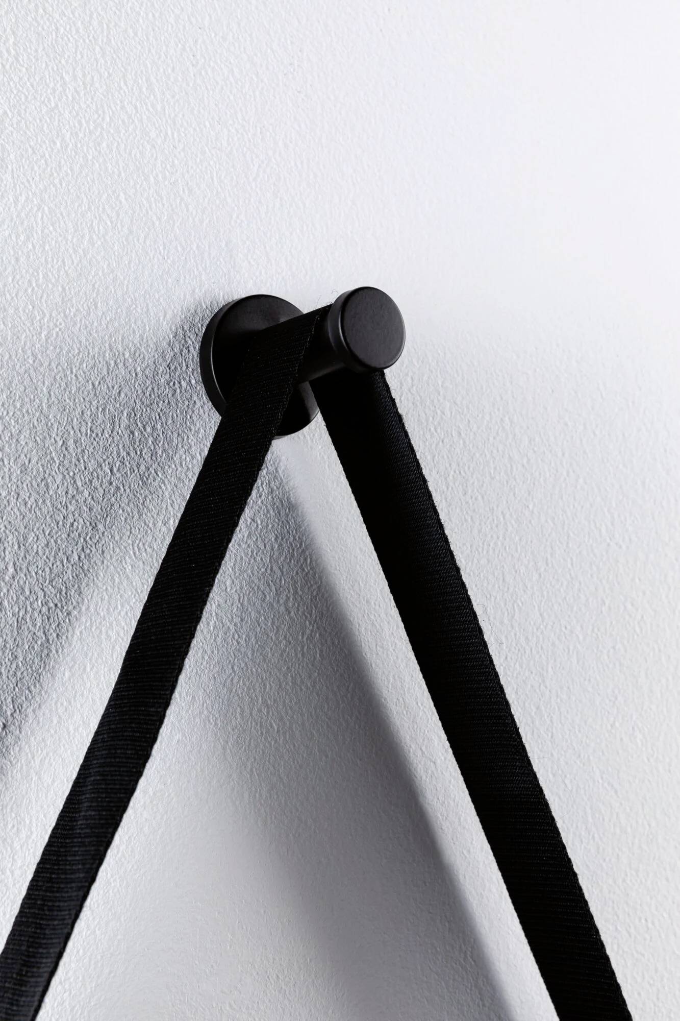 Schwarzer, runder Wandhaken mit schwarzem Band, das an ihm hängt. Minimalistisches Design auf weißem Hintergrund.