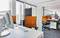 Ein modernes Büro mit weißen Tischen, orangefarbenen Bürostühlen und Trennwänden. Tageslicht fällt durch große Fenster.