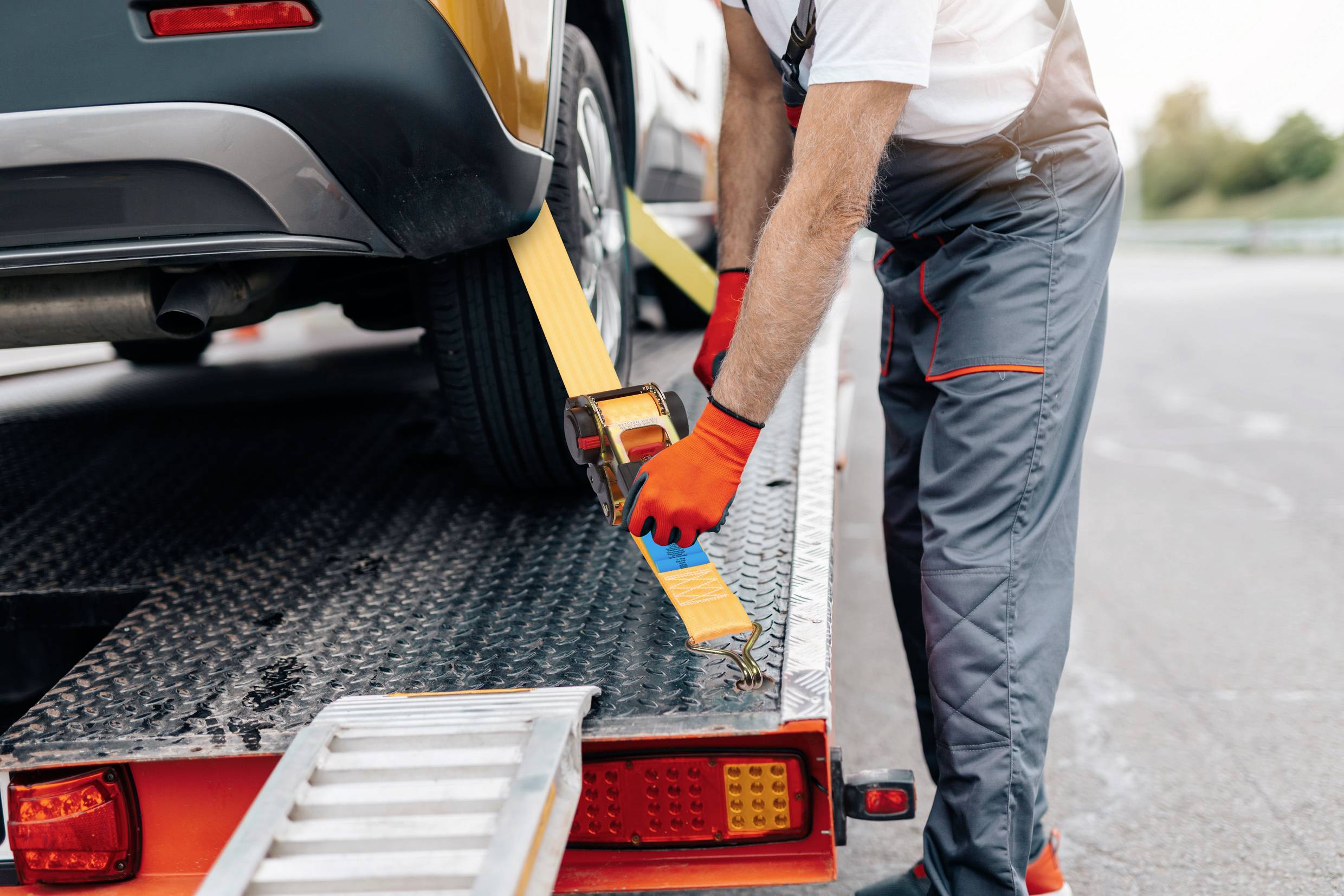Eine Person sichert ein Auto mit einem gelben Gurt auf einem Abschleppwagen. Der Abschleppwagen steht auf einem Parkplatz.
