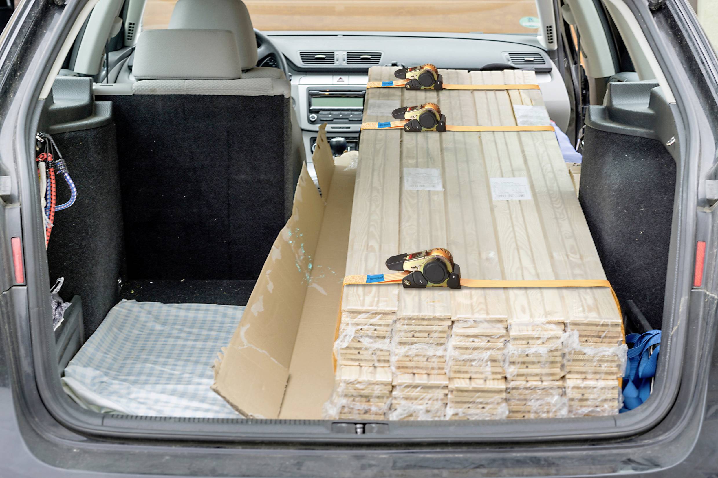 Ein Auto ist von hinten zu sehen, mit einem offenen Kofferraum. Im Kofferraum befinden sich gestapelte Holzbretter, gesichert mit Gurten.