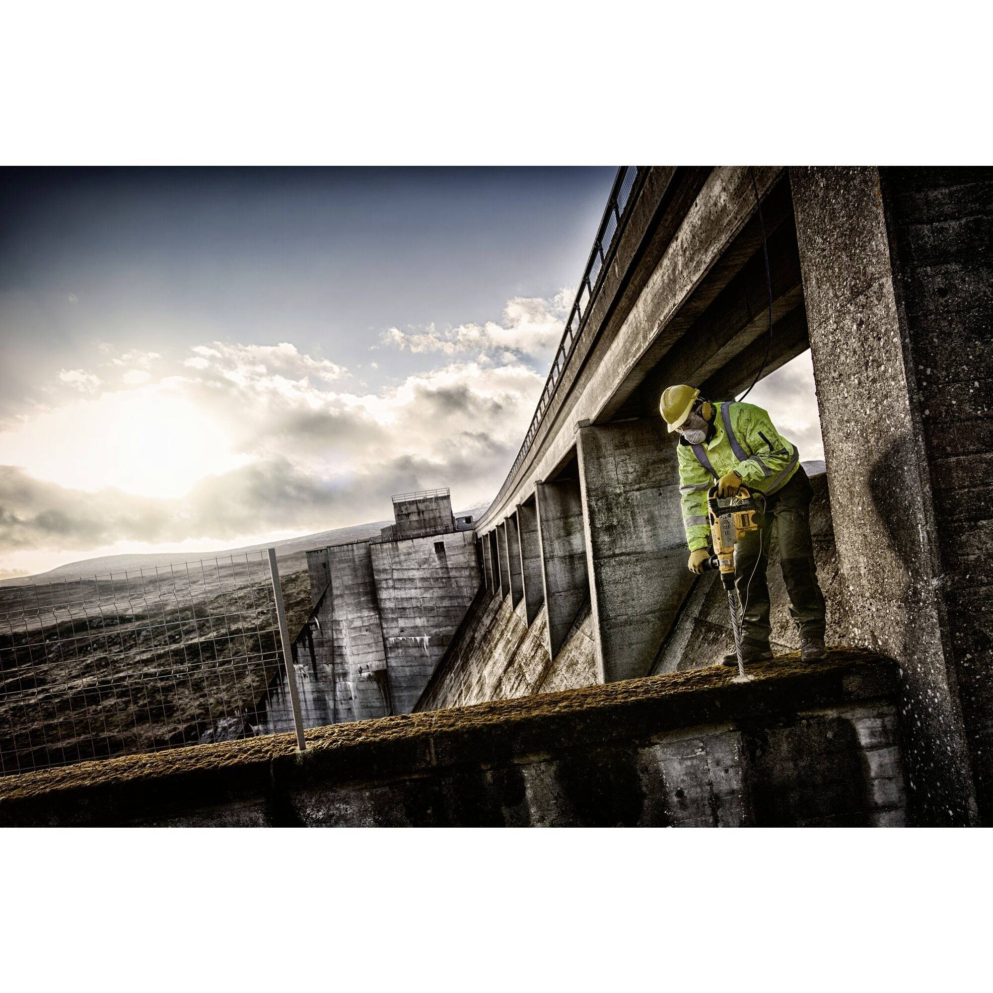 Ein Bauarbeiter in Warnkleidung benutzt einen Presslufthammer auf einer Brücke bei Sonnenuntergang, die Baustelle wirkt groß und industriell.