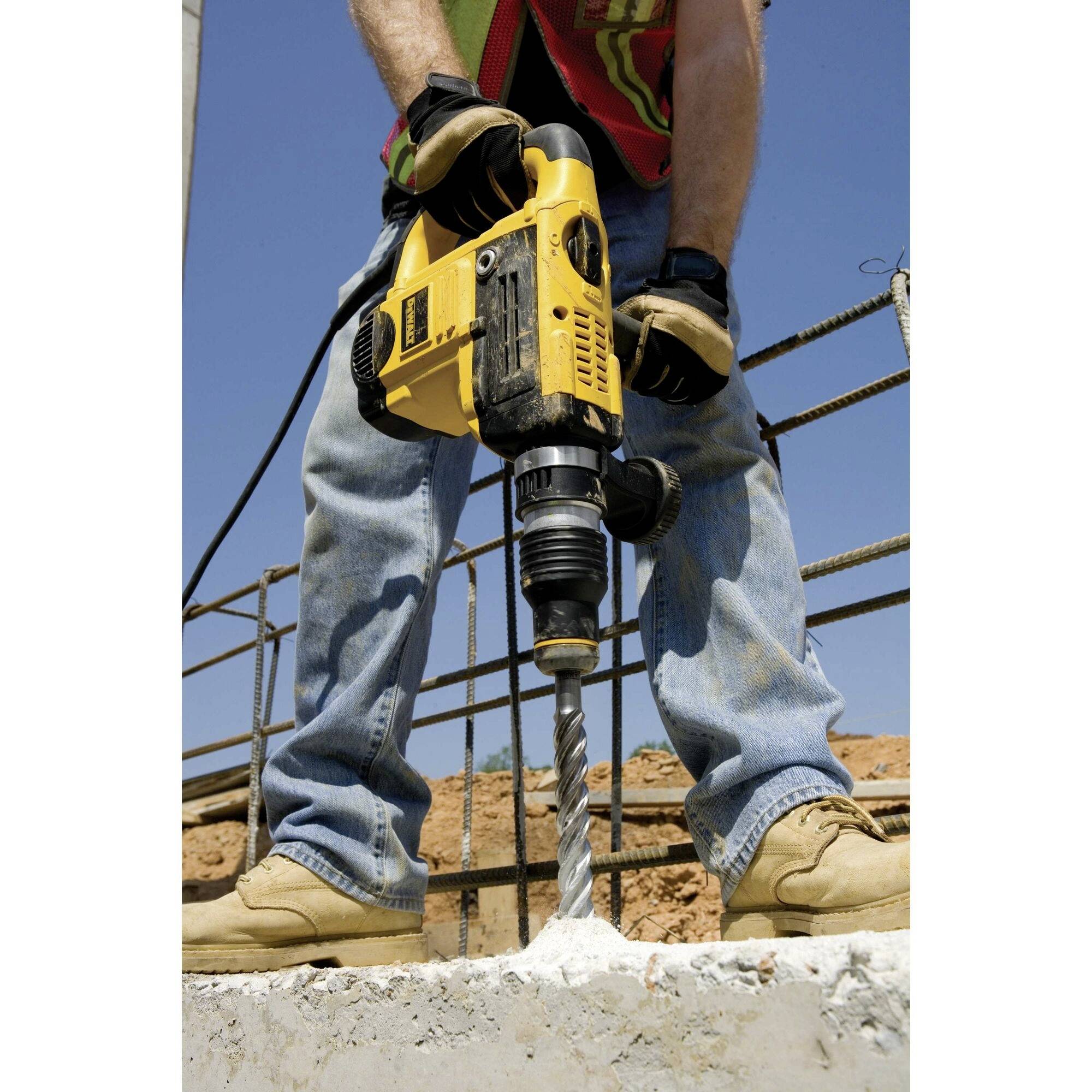 Eine Person bohrt mit einer gelben Bohrmaschine ein Loch in Beton; sie trägt Arbeitskleidung und Arbeitshandschuhe.