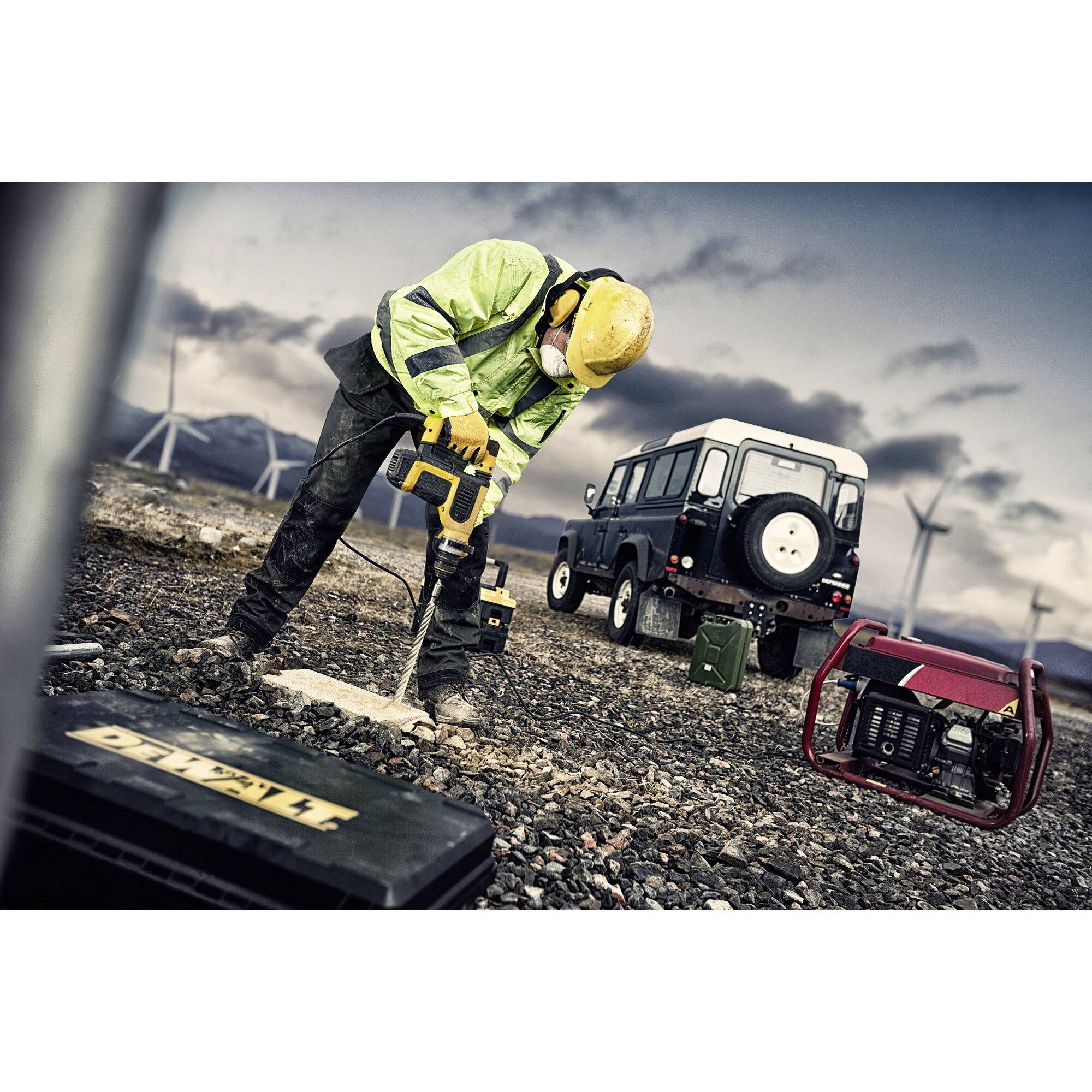 Ein Bauarbeiter mit Schutzkleidung bohrt in den Boden, während ein Geländefahrzeug und ein Generator im Hintergrund zu sehen sind.