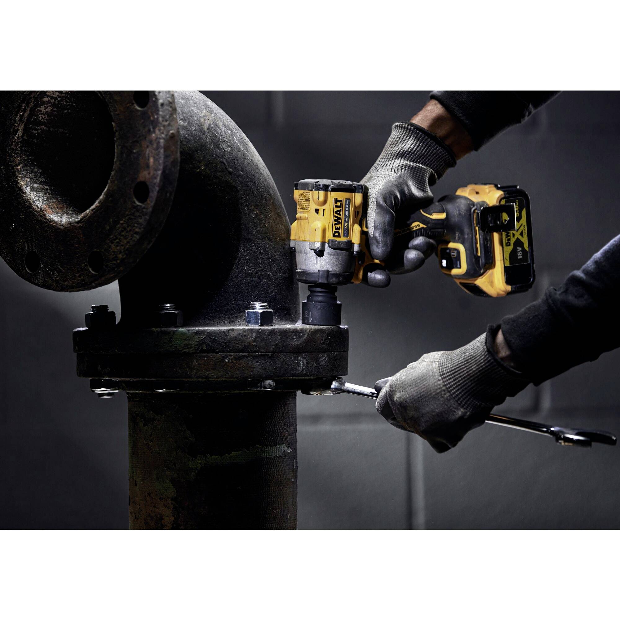 'Zwei Hände in Arbeitshandschuhen verwenden Bohrmaschine und Schraubenschlüssel, um Schraube an großem Metallrohr zu fixieren.'