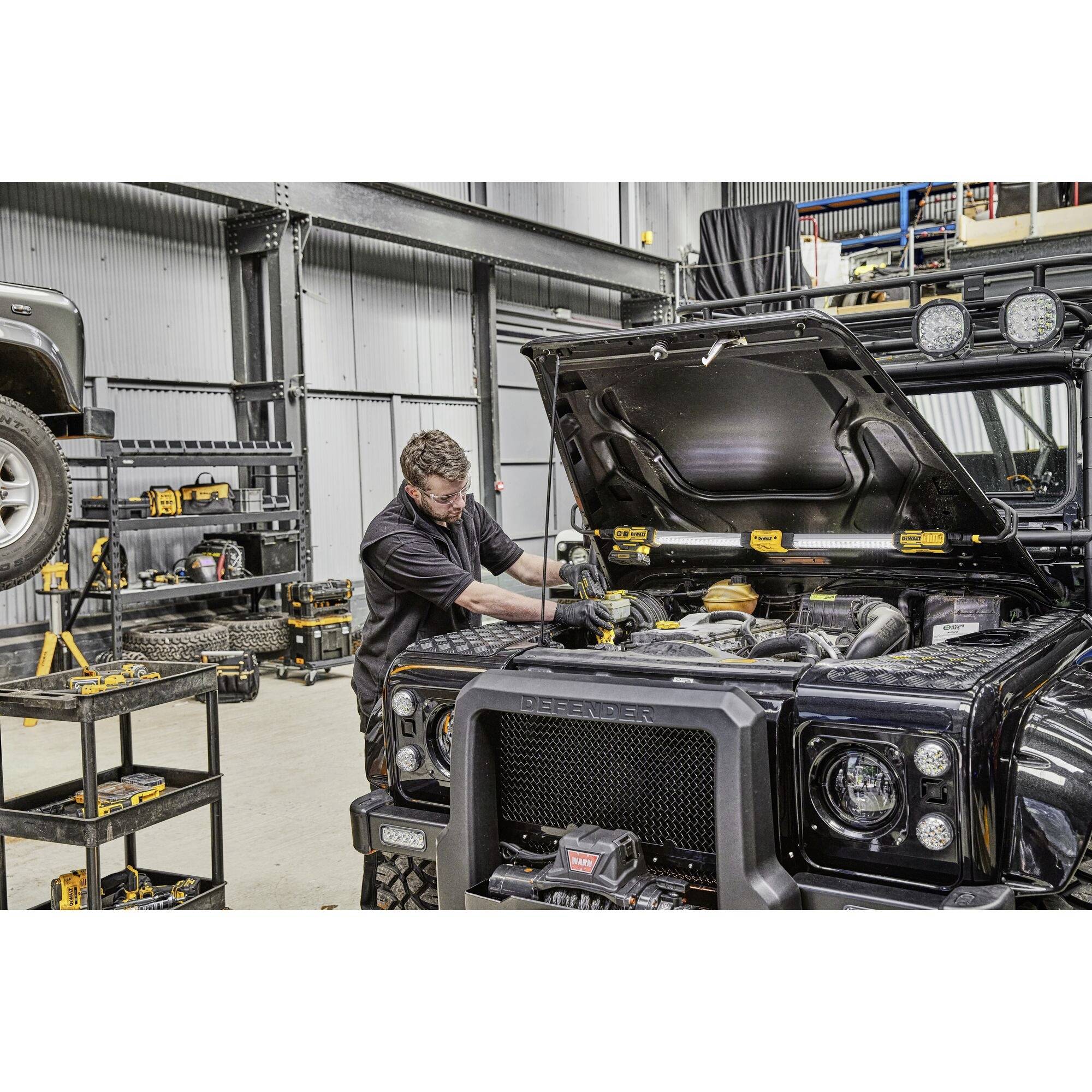 Mechaniker arbeitet in einer Werkstatt an einem schwarzen Geländewagen mit geöffneter Motorhaube. Werkzeuge sichtbar im Hintergrund.