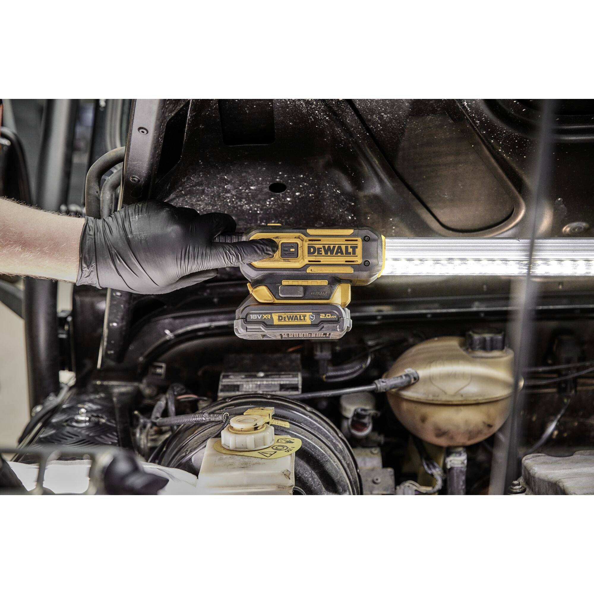 Eine Person mit schwarzen Handschuhen hält ein DeWalt-Werkzeug unter die Motorhaube eines Fahrzeugs, um den Motorraum zu beleuchten.