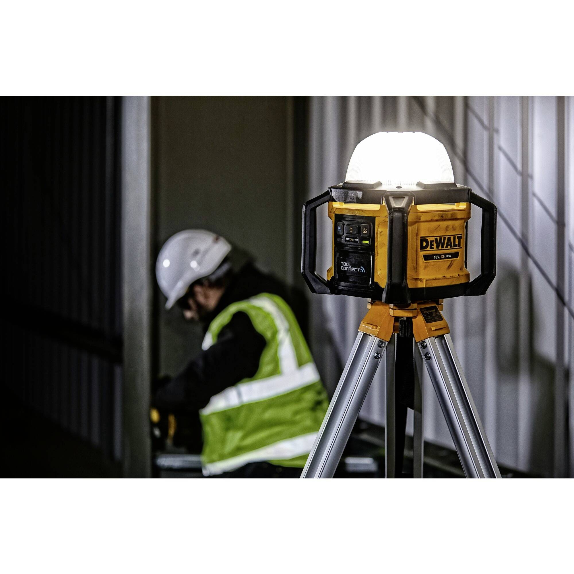 Ein Arbeiter in Schutzkleidung installiert eine Metallrahmenkonstruktion, während rechts ein DeWalt-Baustrahler auf einem Stativ leuchtet.
