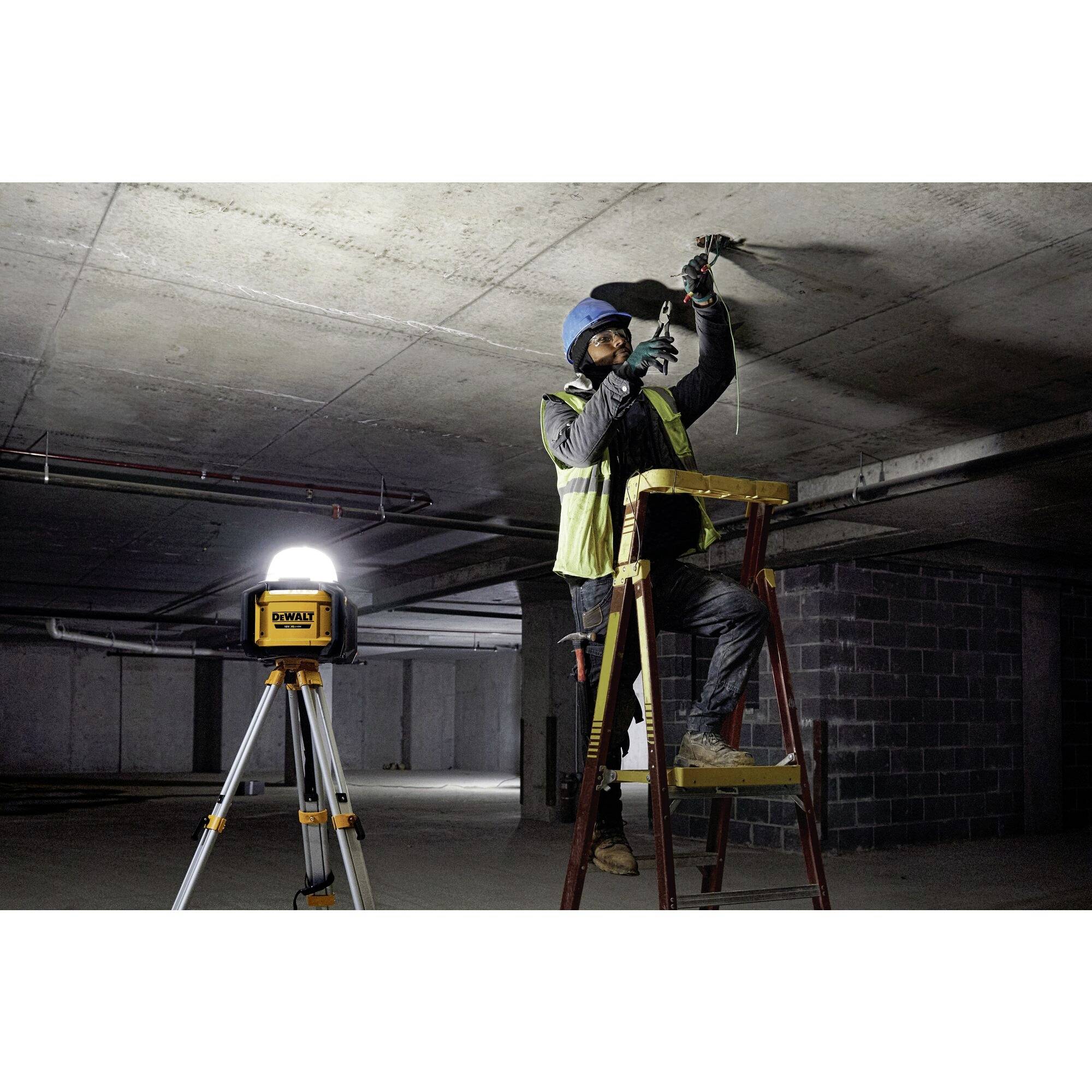 Ein Bauarbeiter in Sicherheitskleidung steht auf einer Leiter und arbeitet an der Decke eines Parkhauses. Eine Lampe leuchtet den Bereich aus.