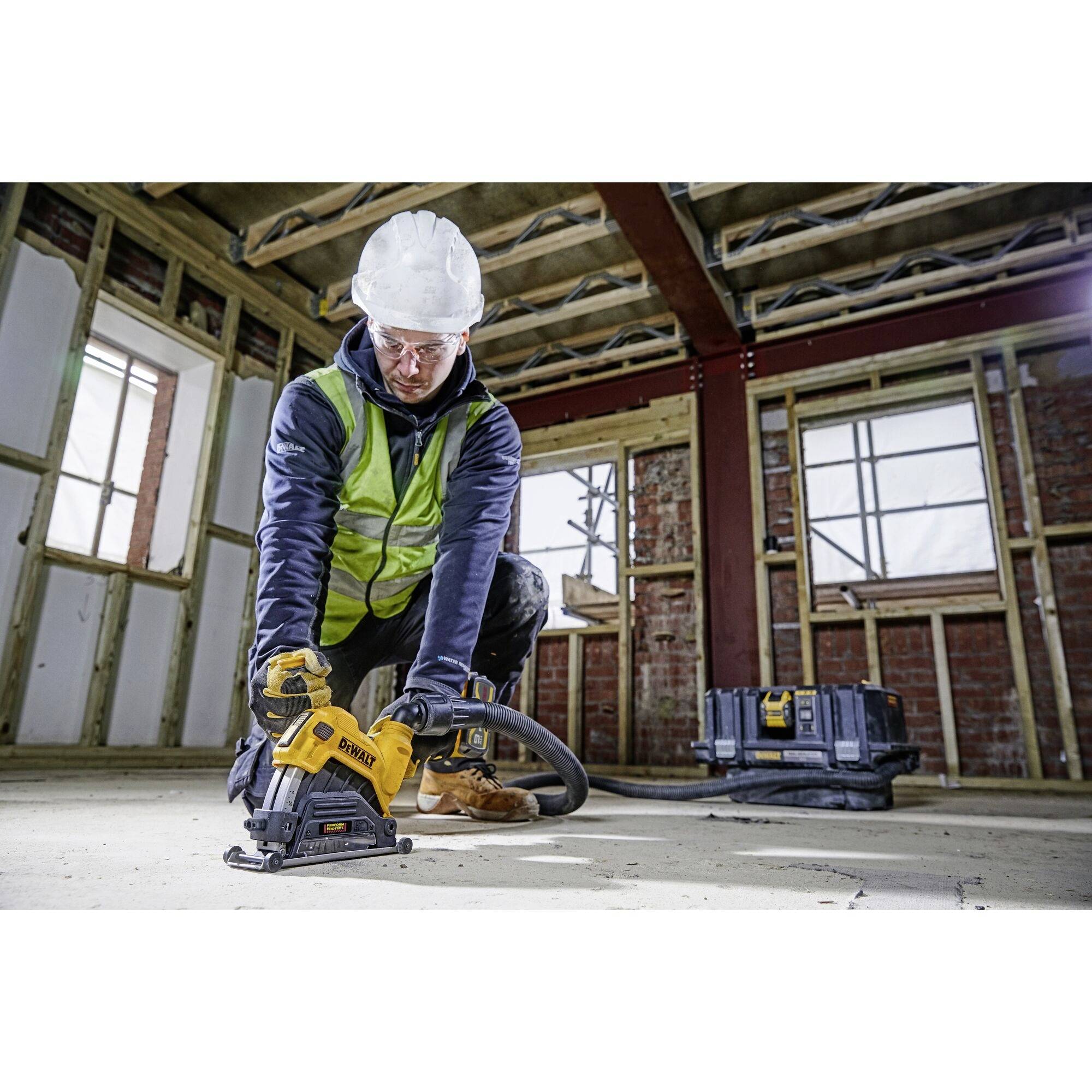 Eine Person in Schutzkleidung arbeitet mit einem Elektrowerkzeug auf einer Baustelle. Sie trägt einen weißen Helm und eine Schutzweste.