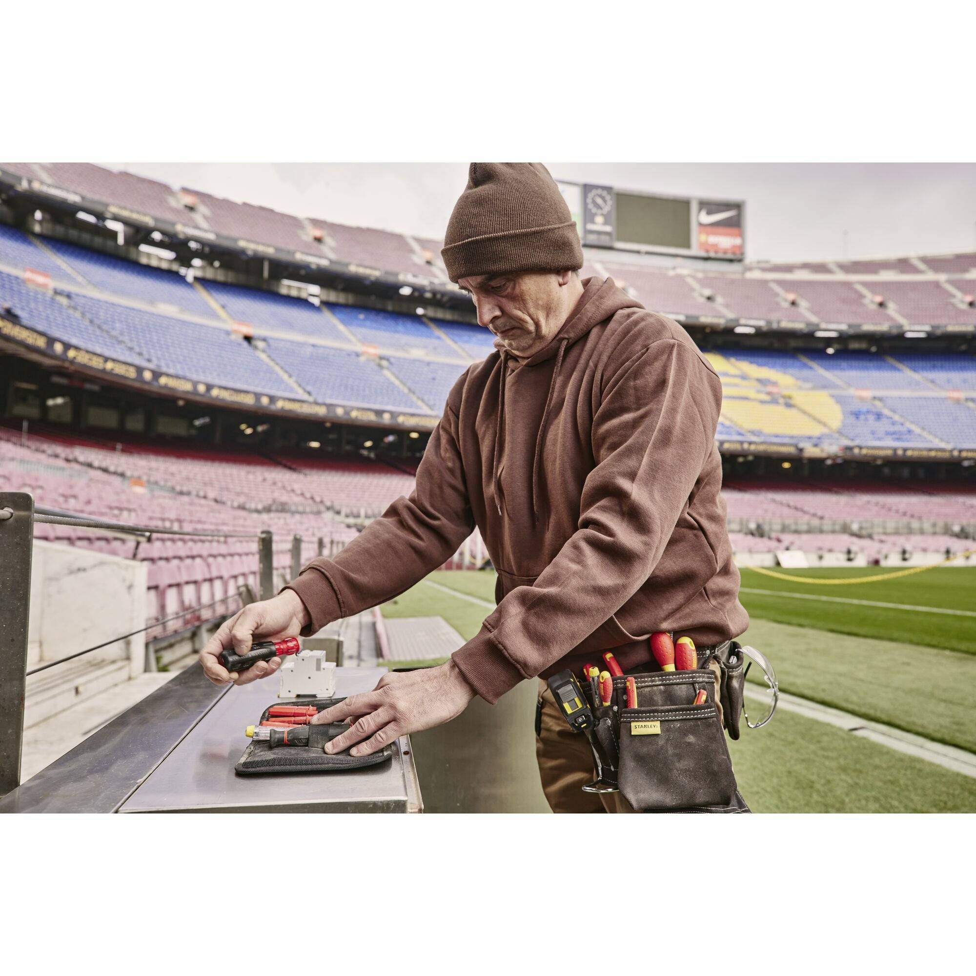 Eine Person in Arbeitskleidung repariert Elektronik in einem leeren Stadion. Sie trägt Werkzeuge in einer Tasche an der Hüfte.