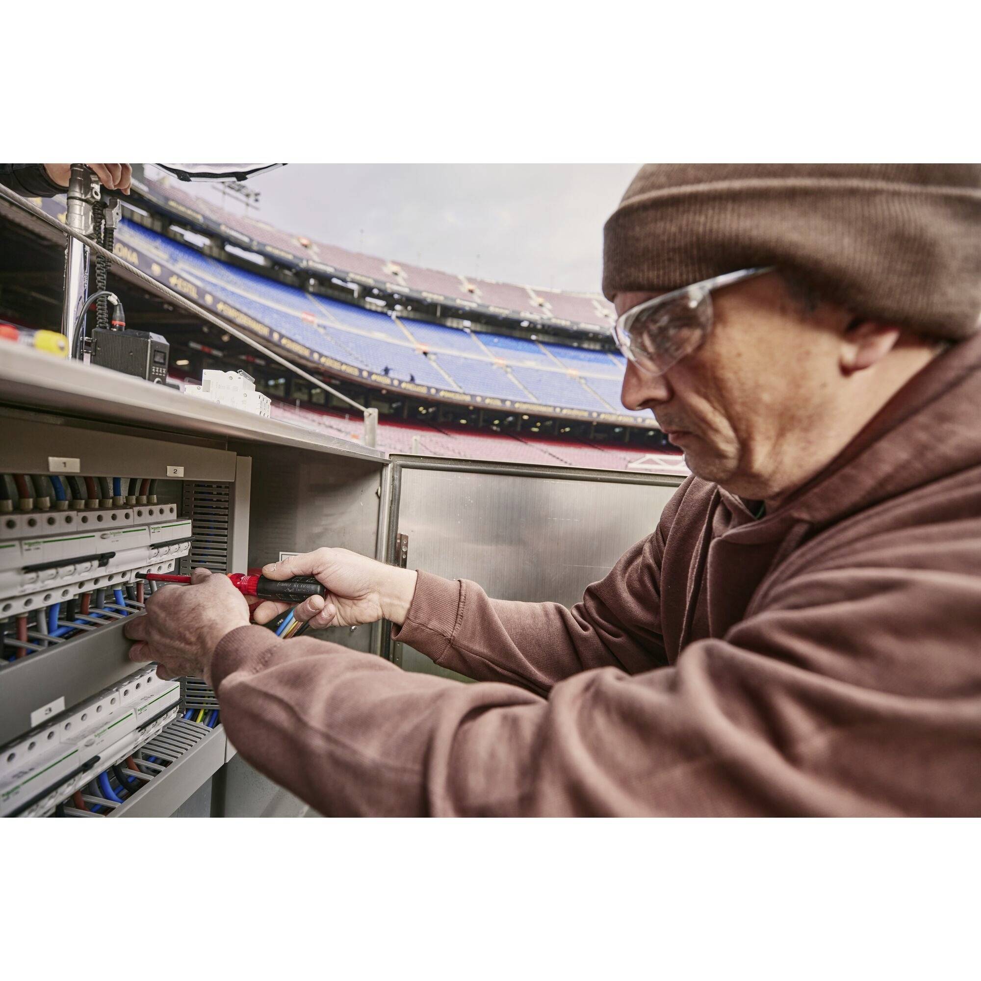 Ein Mann in Schutzbrille arbeitet an einem Schaltschrank in einem Stadion. Er hält ein Werkzeug und justiert Kabel. Im Hintergrund sind leere Tribünen zu sehen.