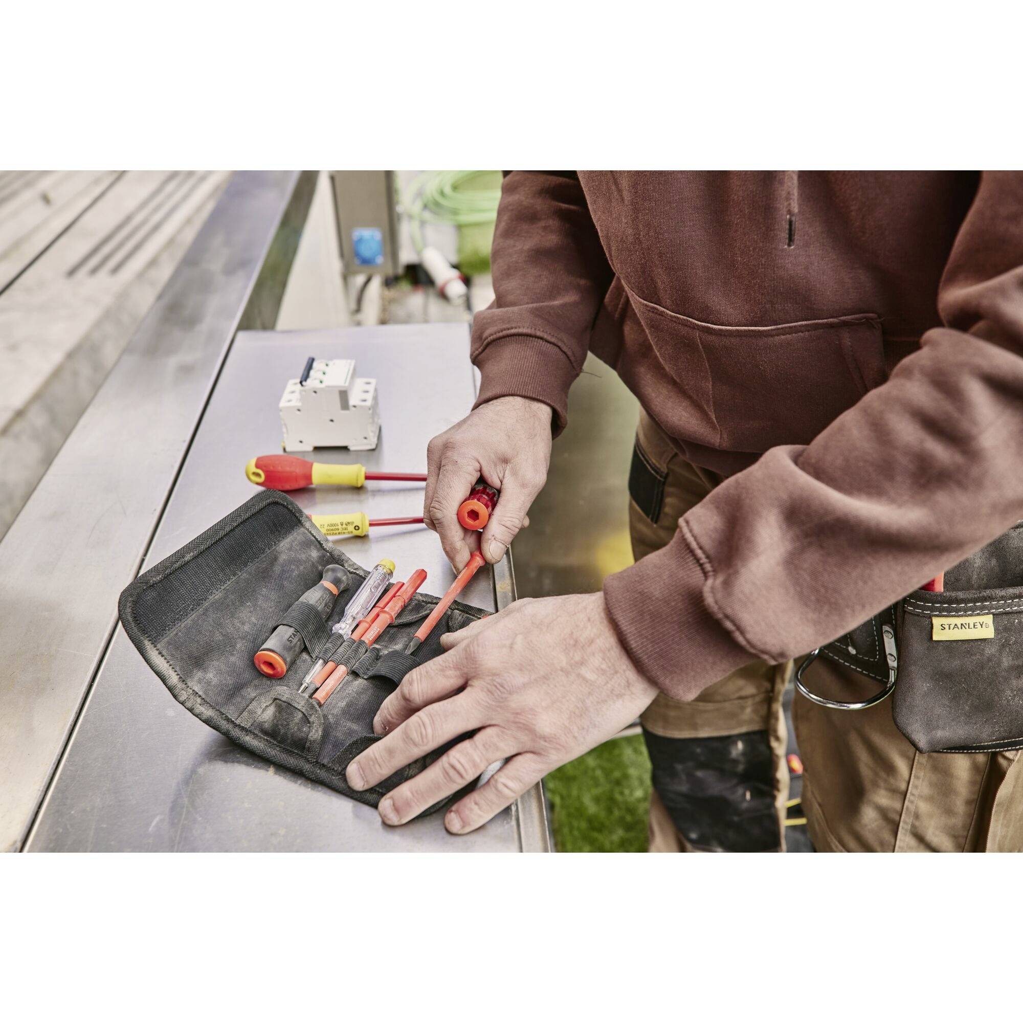 Eine Person repariert elektrische Geräte mit Werkzeugen auf einem Tisch. Schraubendreher und Sicherungskasten sind sichtbar.