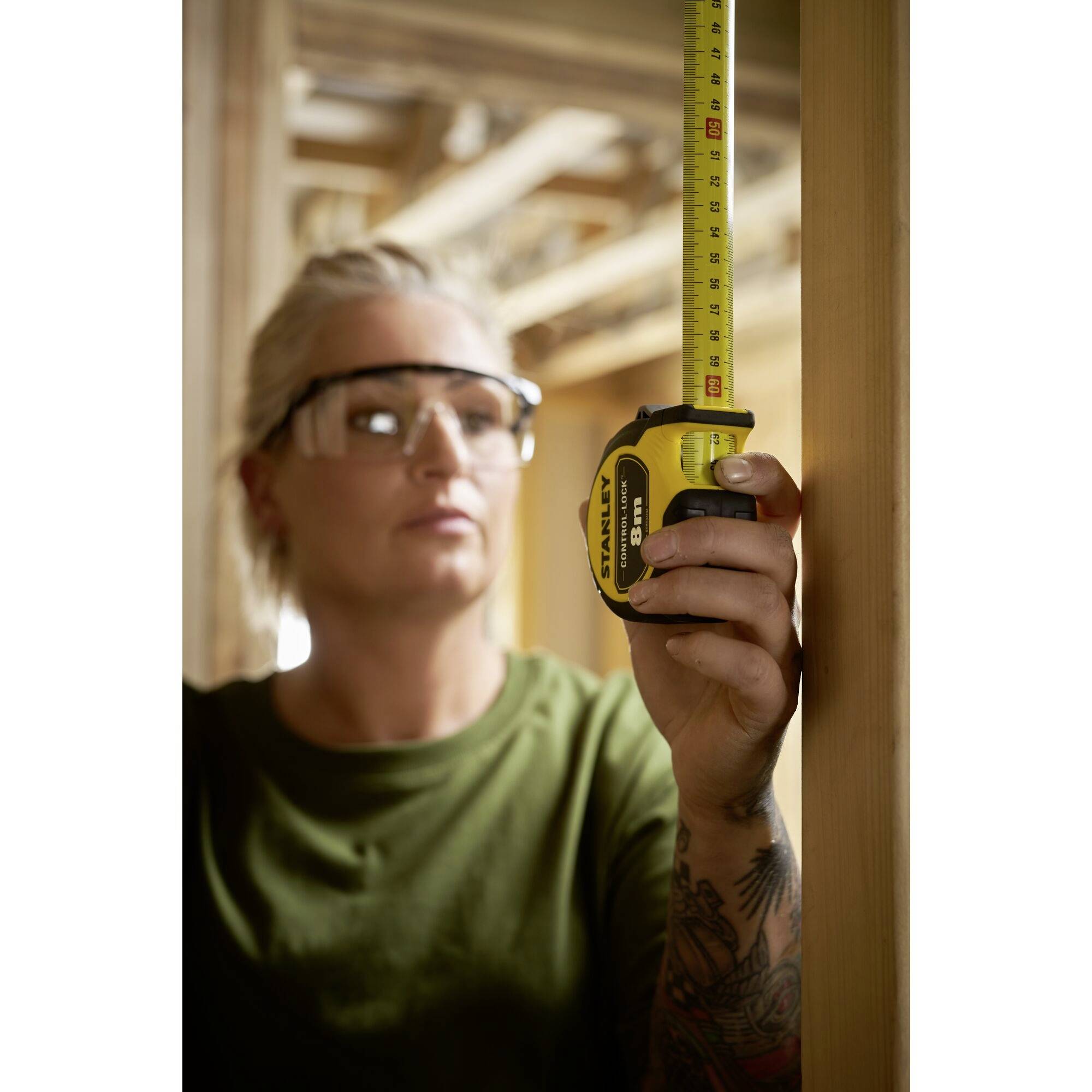 Eine Person misst ein Holzstück mit einem Maßband in einer Baustelle. Die Person trägt Schutzbrille und hat tätowierte Arme.