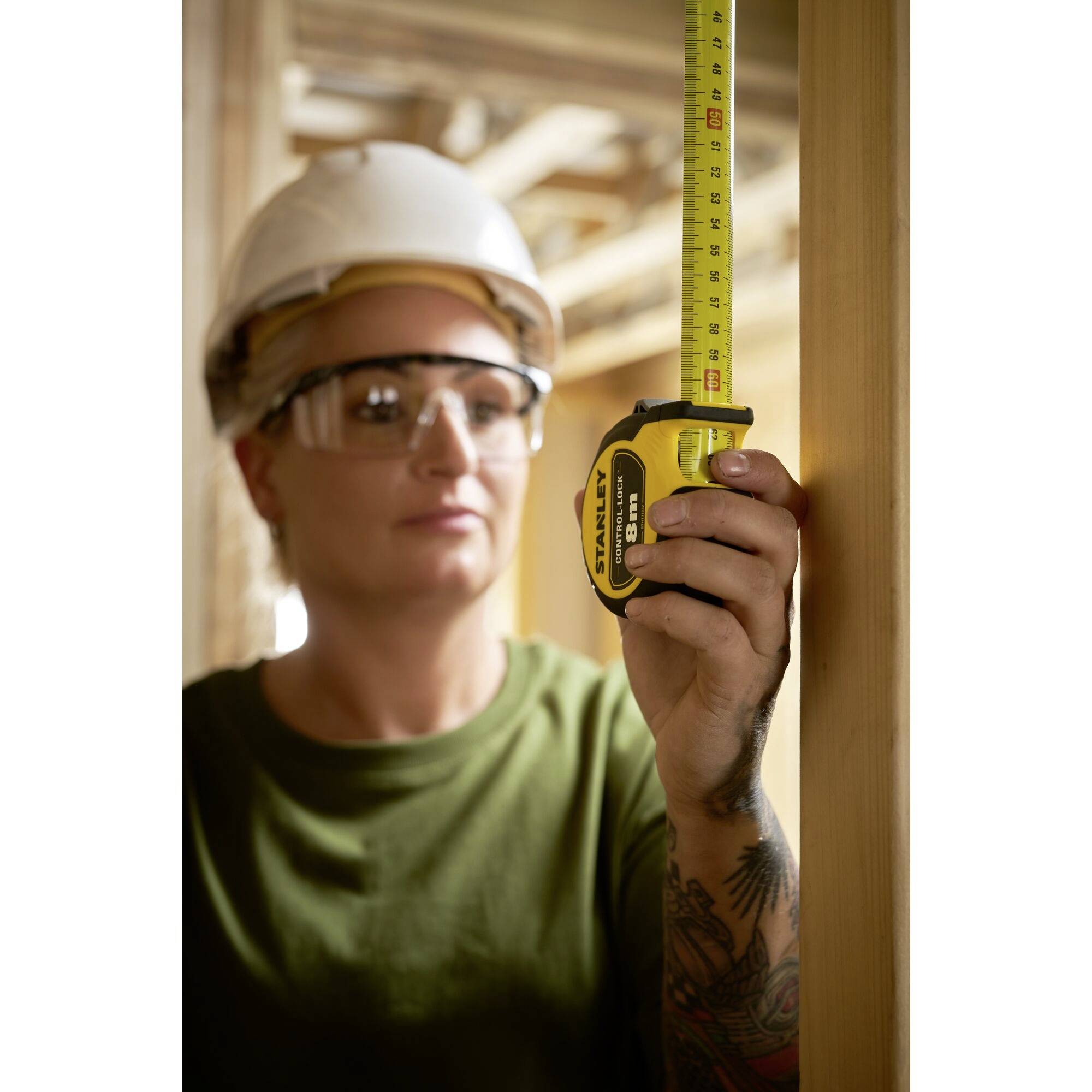 Eine Person mit Schutzhelm und Schutzbrille misst mit einem Maßband die Höhe eines Holzpfostens in einer Baustellenumgebung.