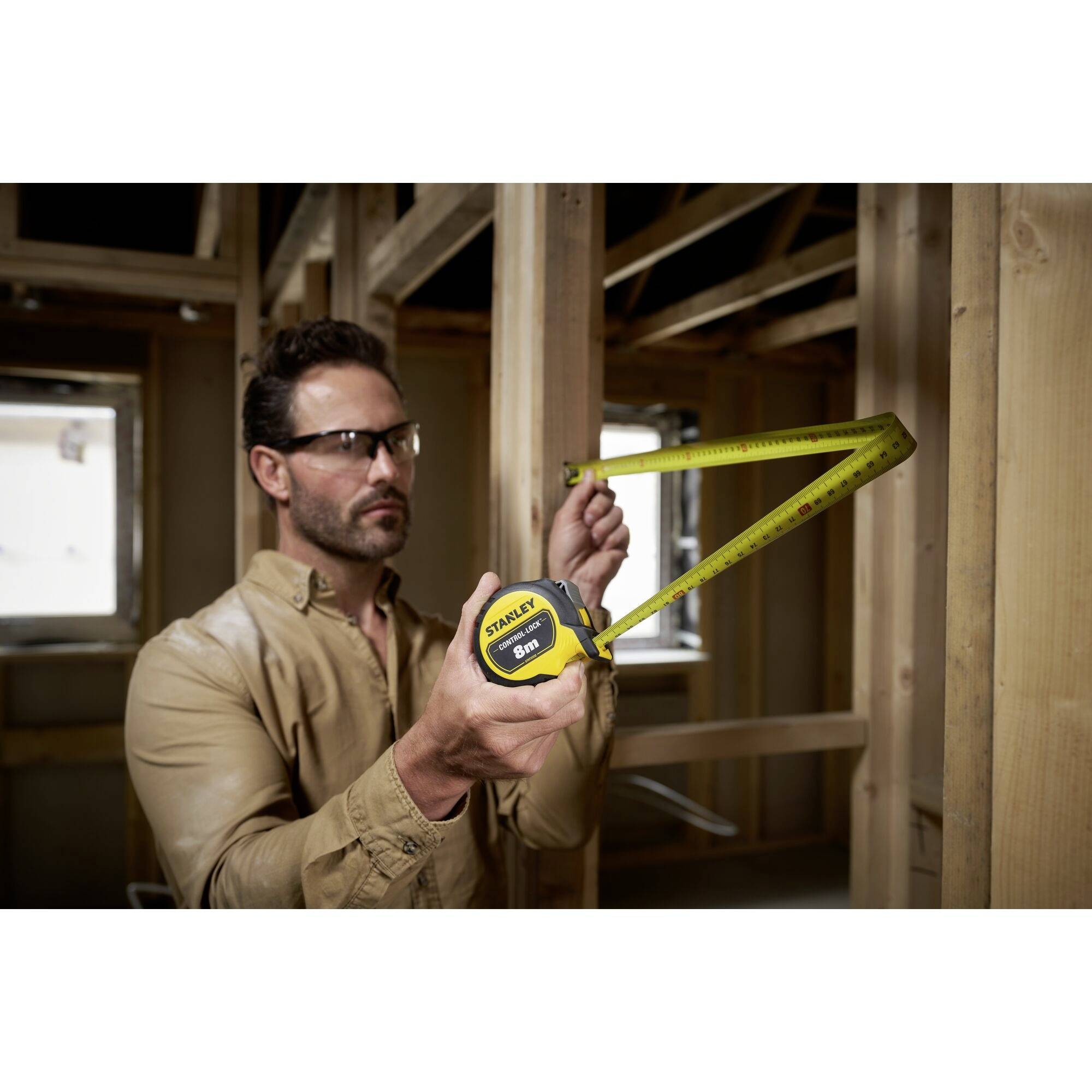 Eine Person misst einen Holzbalkenrahmen mit einem Maßband in einem im Bau befindlichen Raum. Sie trägt Schutzbrille.