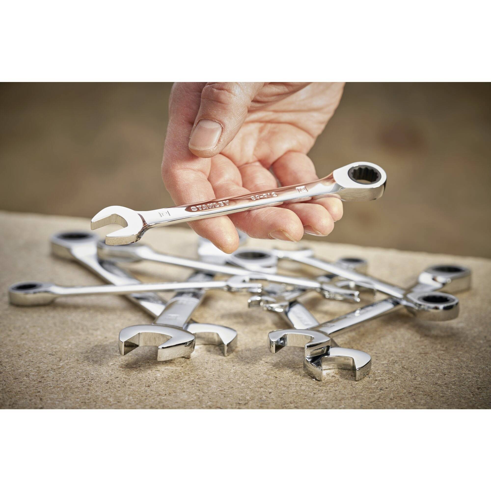 Eine Hand hält einen Ringschlüssel, umgeben von mehreren auf einem Tisch liegenden Schraubenschlüsseln.