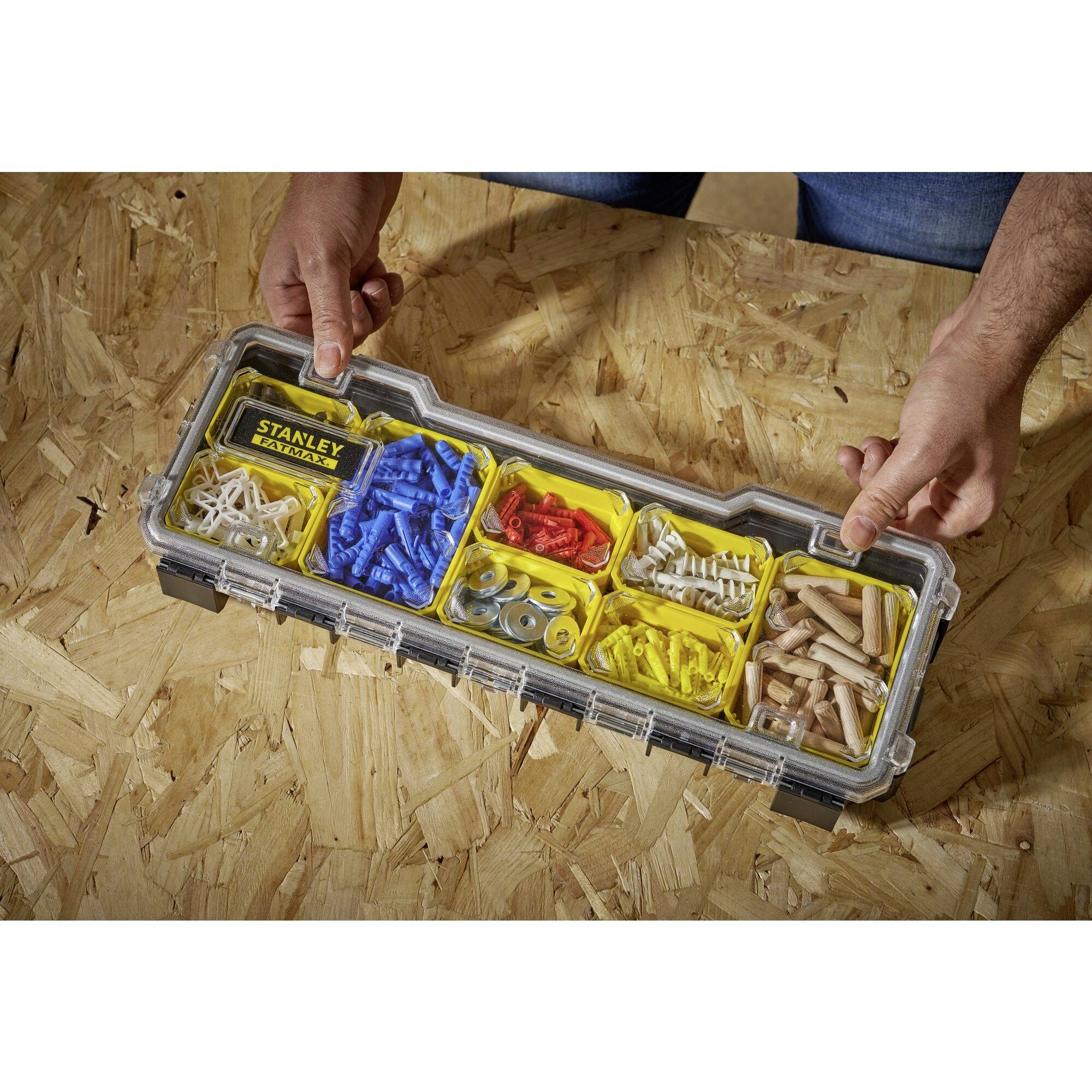 Eine Person hält eine geöffnete Aufbewahrungsbox mit verschiedenen Schrauben, Dübeln und Befestigungsmaterialien auf einem Holztisch.