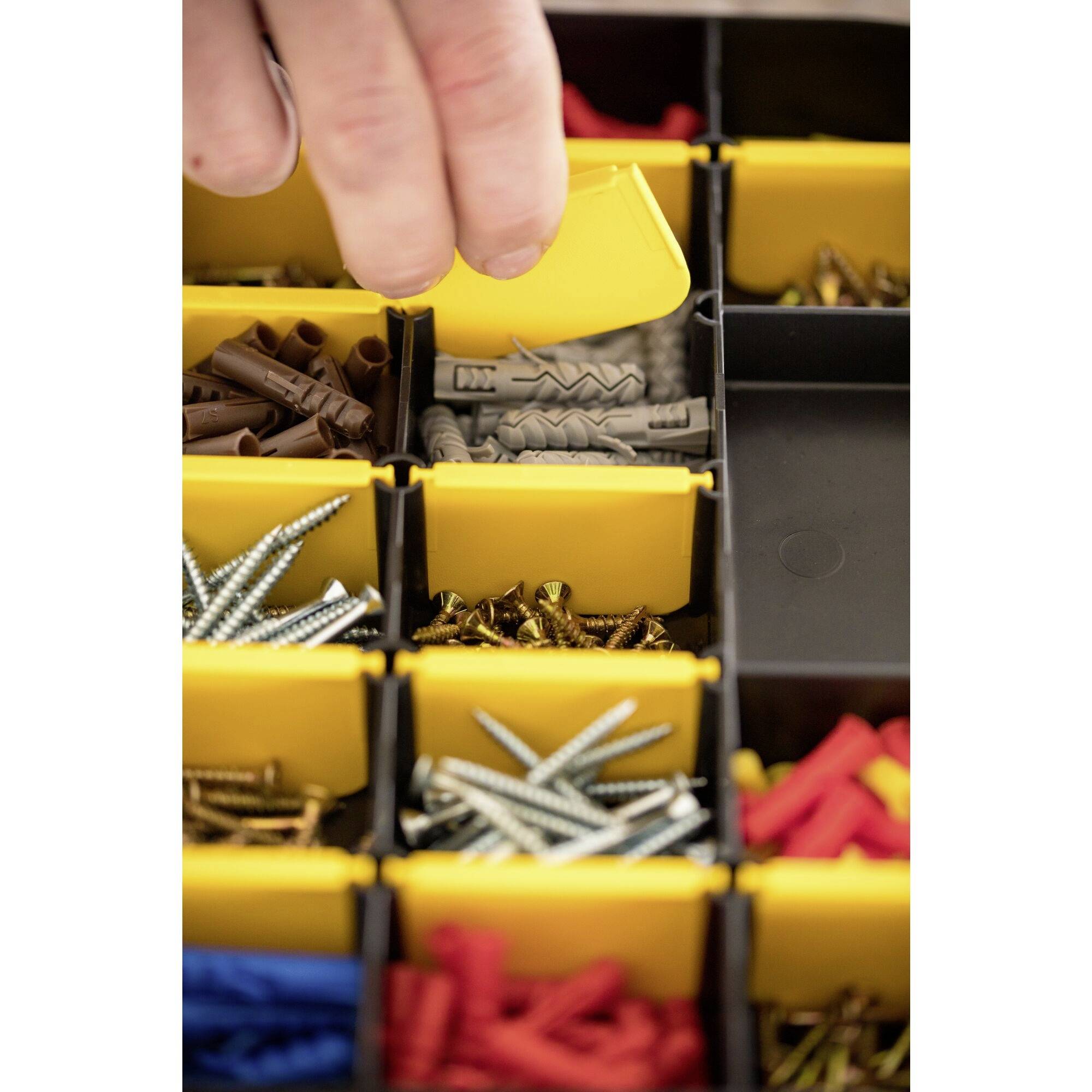 Eine Hand entnimmt eine graue Schraubendübel aus einem gelben Fach einer Sortierbox, die verschiedene Schrauben und Dübel enthält.