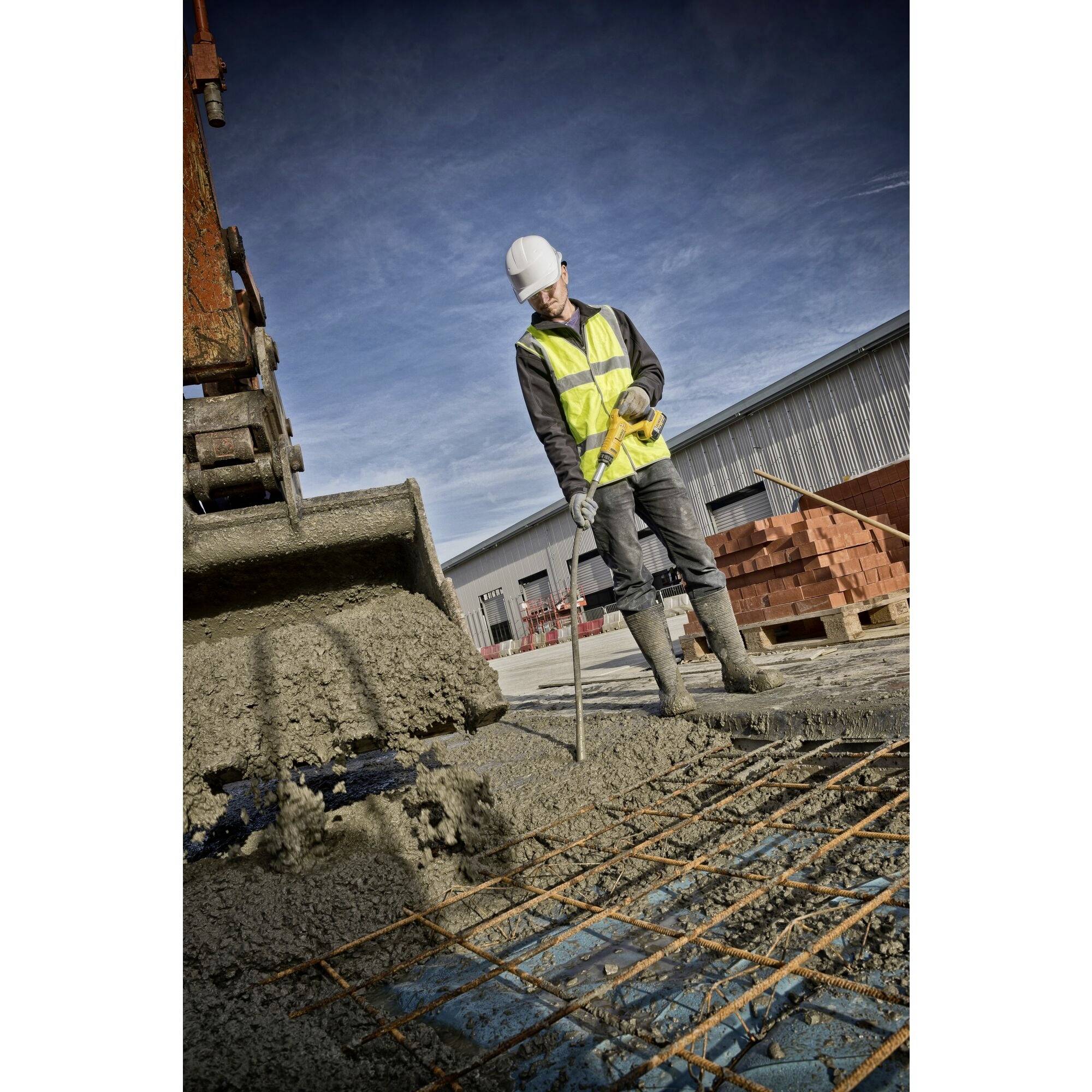 Ein Bauarbeiter in Schutzkleidung verteilt Beton mit einem Werkzeug neben einem Bagger auf einer Baustelle mit Ziegelstapeln im Hintergrund.