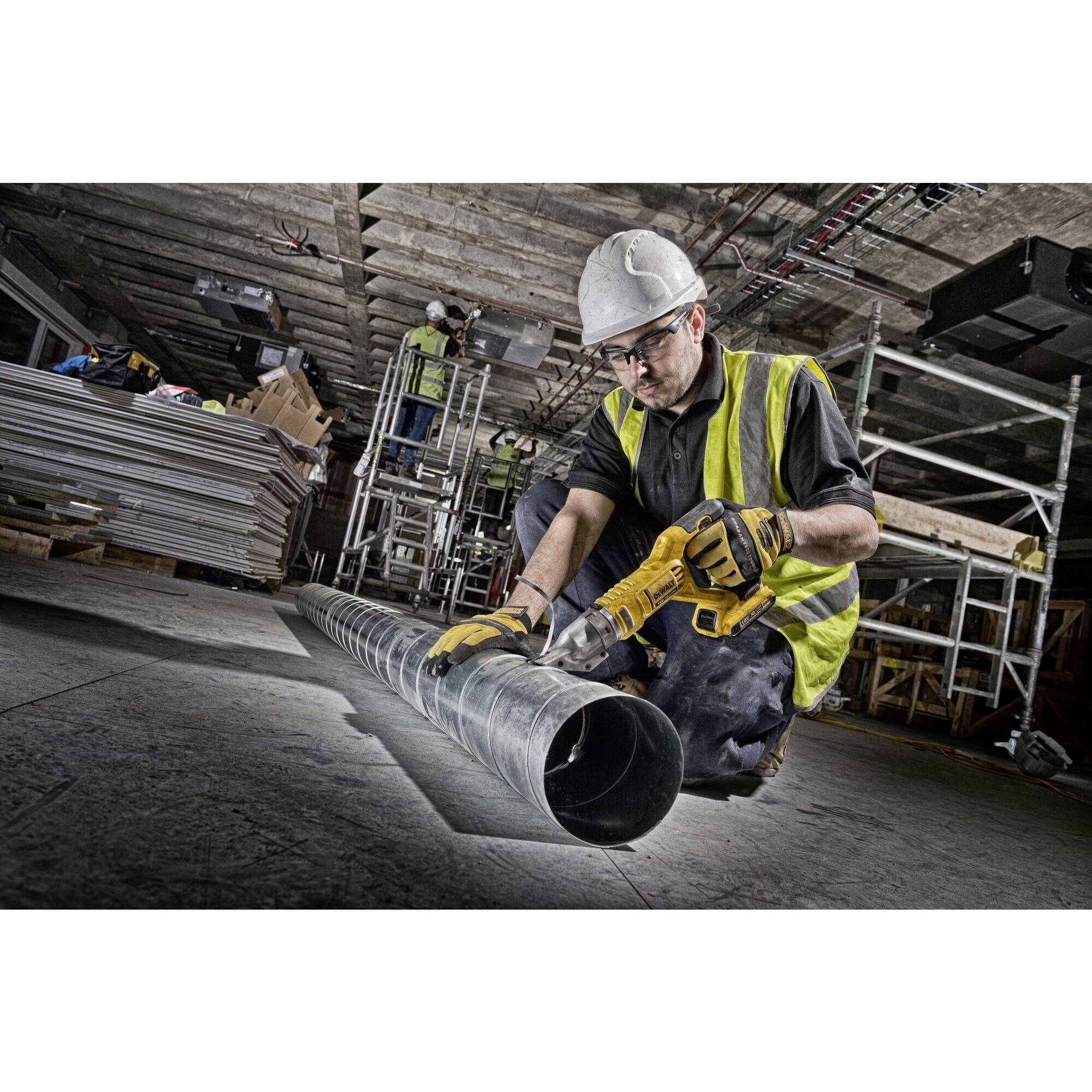 'Arbeiter mit Schutzkleidung schneidet Metallrohr auf Baustelle. Hintergrund zeigt Gerüste und Baumaterialien.'