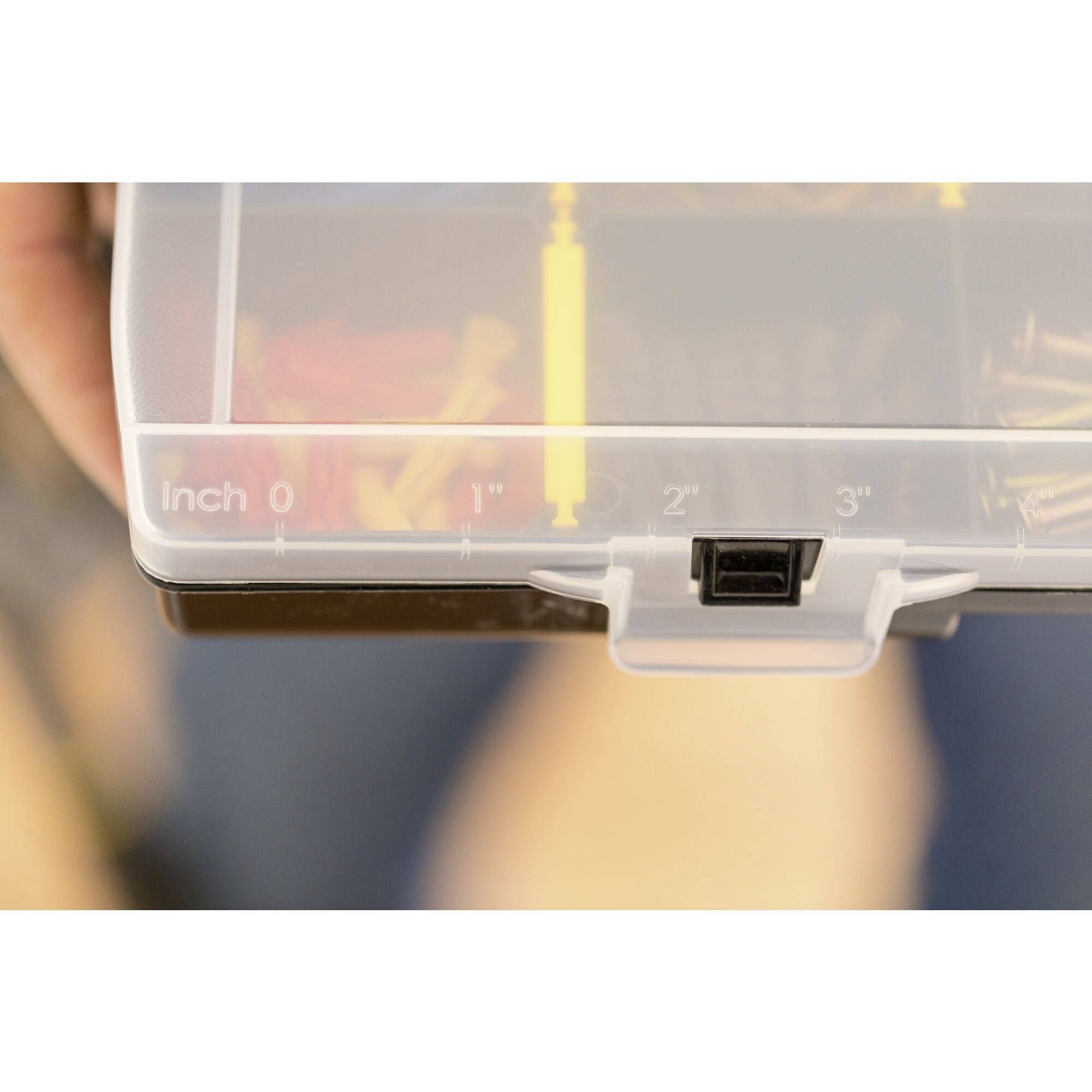 Hand hält durchsichtige Aufbewahrungsbox mit gelbem Maßband. Maßangaben in Zoll (0, 1, 2, 3) auf der Box sichtbar.