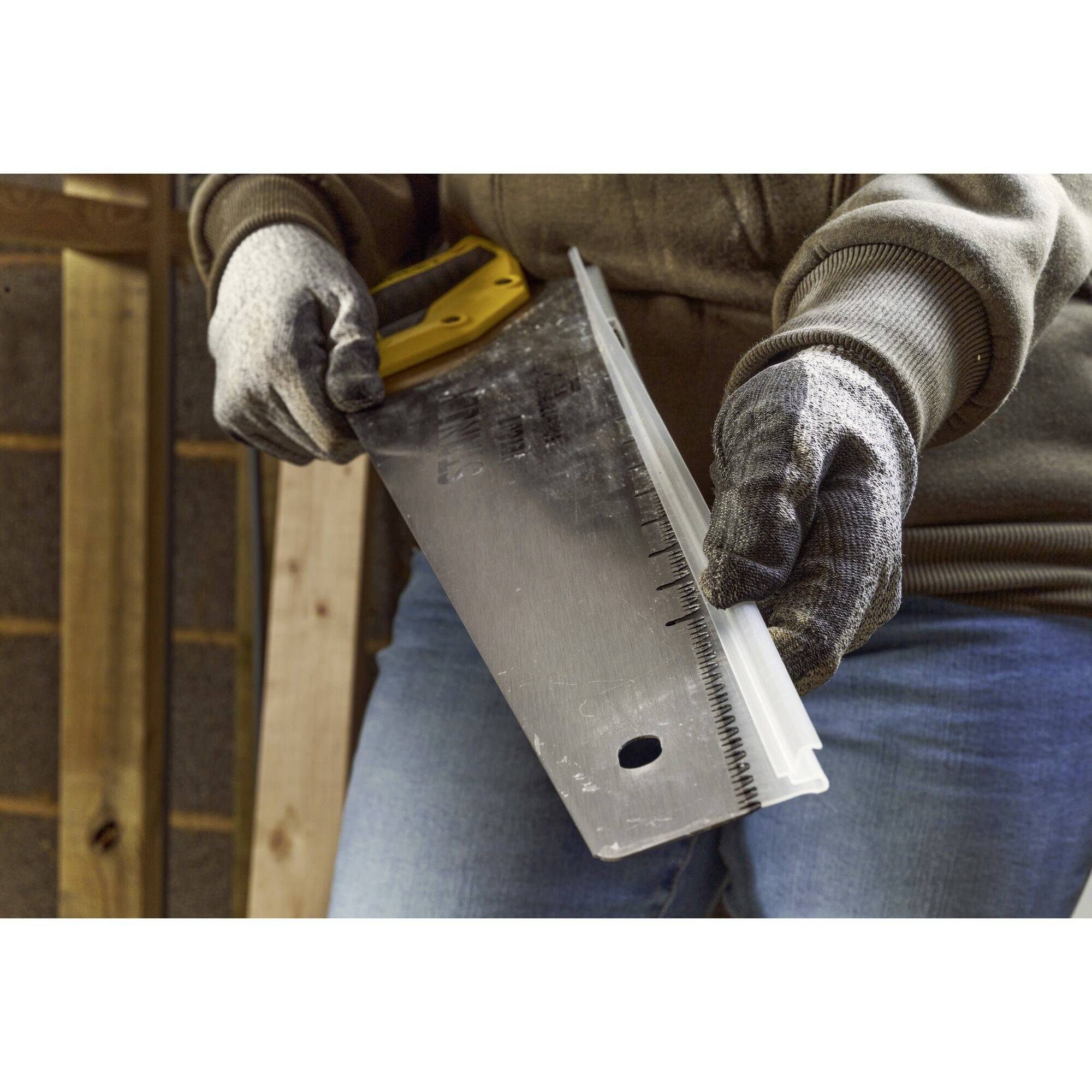 Eine Person hält eine Handsäge mit gelbem Griff und Schutzkleidung. Sie arbeitet in einer Werkstatt oder Baustelle.