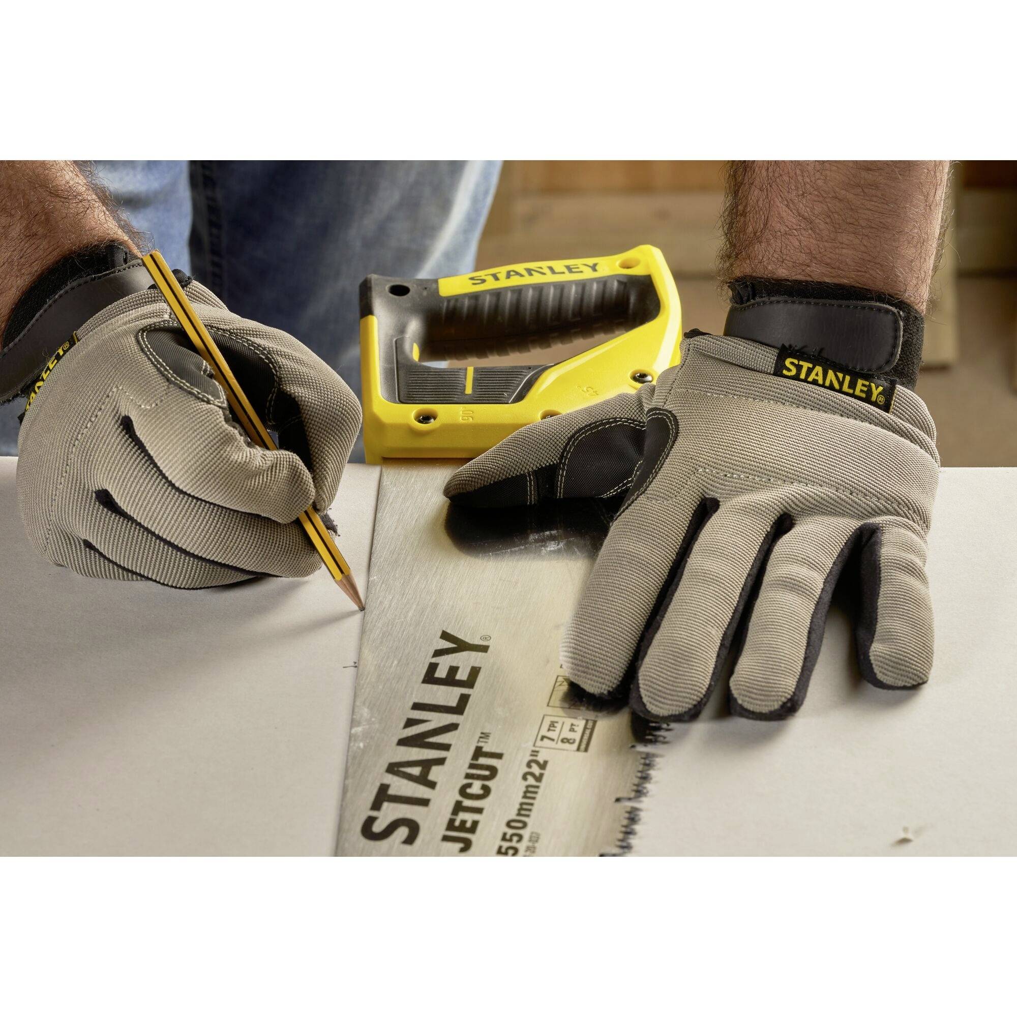 Eine Person markiert mit einem Bleistift eine Linie auf einem Stück Holz, während sie eine Säge und Handschuhe von Stanley trägt.