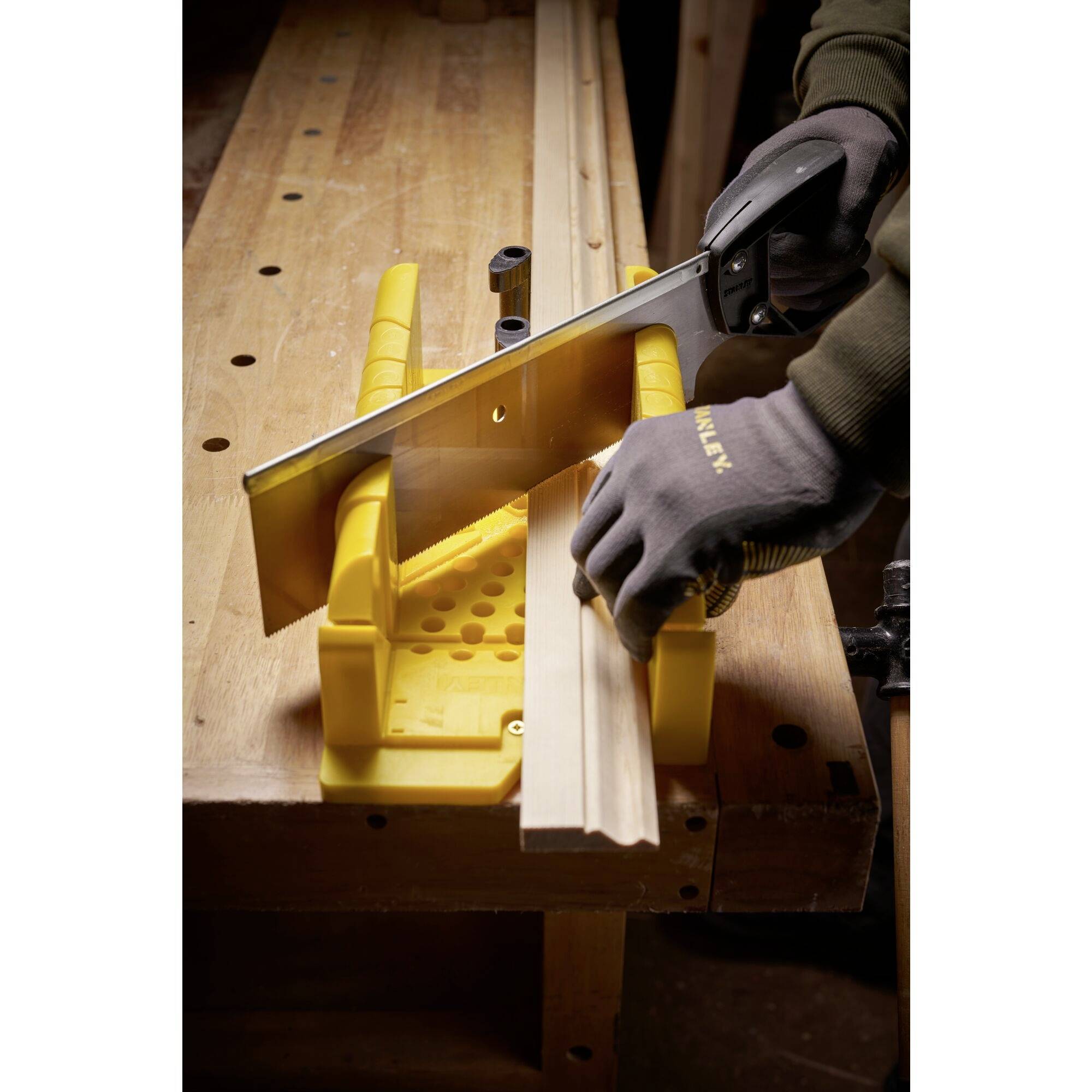 Person sägt ein Stück Holz mit einer Gehrungssäge in einer Werkstatt. Die Hände tragen Arbeitshandschuhe, das Holz ist in einer gelben Sägehilfe eingespannt.