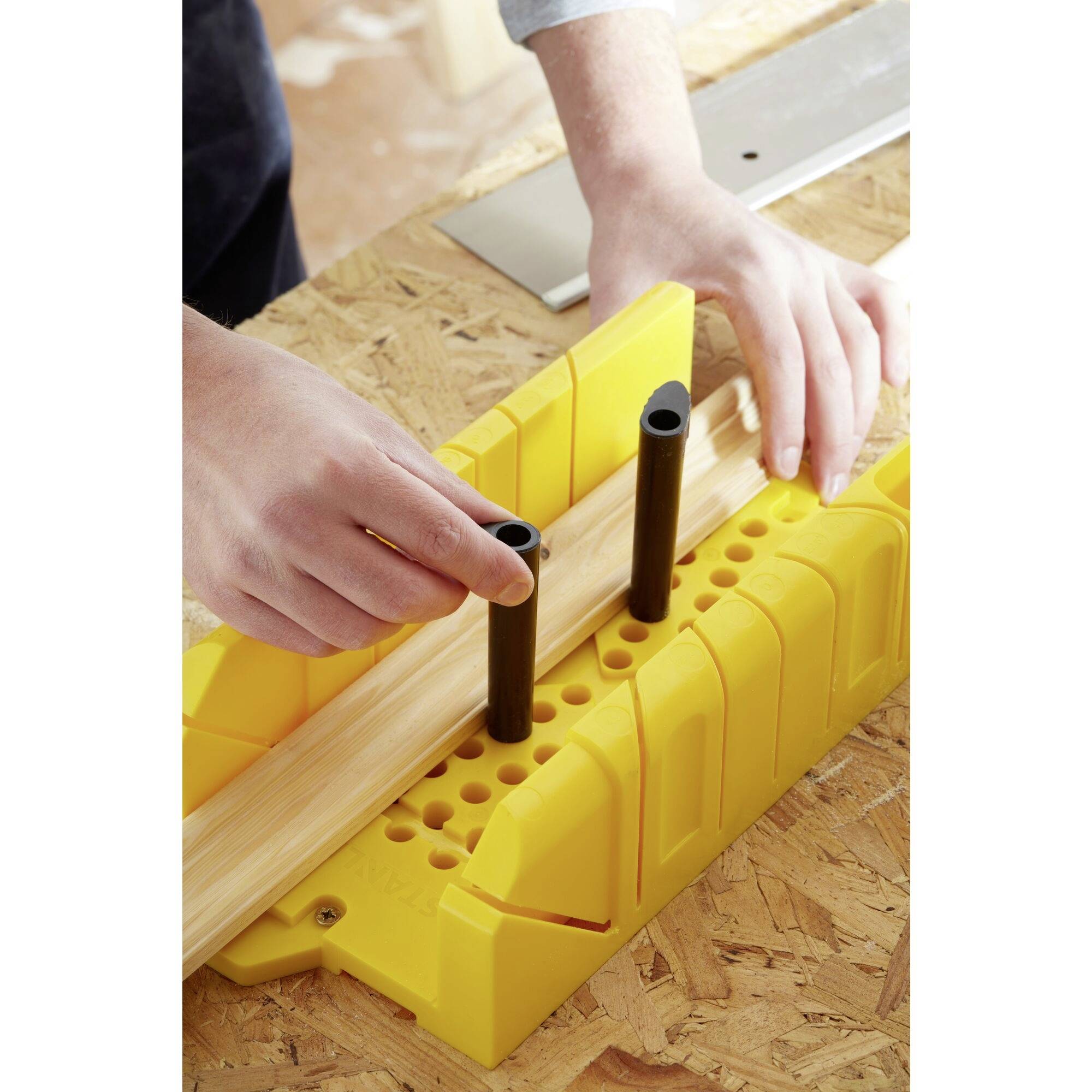 Eine Person richtet ein Holzstück in einer gelben Gehrungslade aus, bereit zum Sägen. Hände halten das Holz fest.
