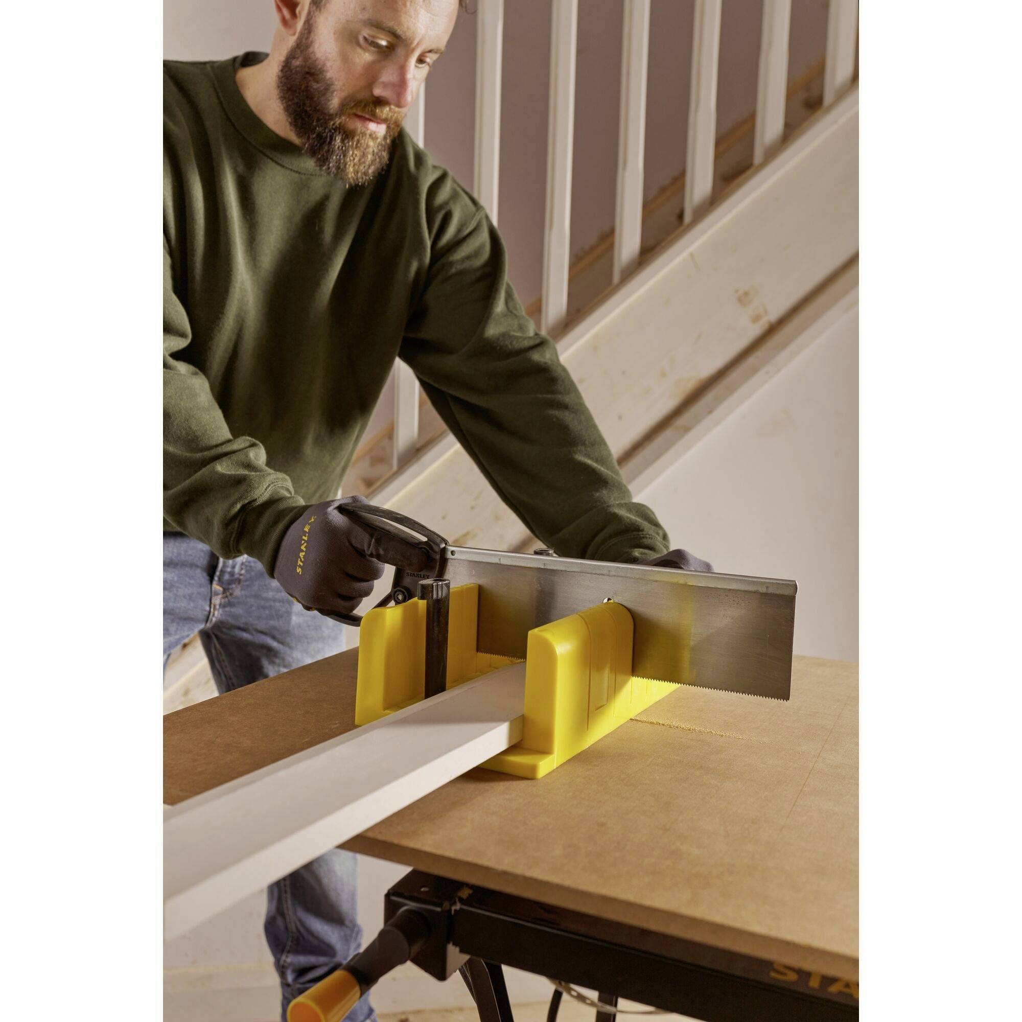 Eine Person schneidet mit einer Handsäge ein Brett auf einer Tischwerkbank. Die Person trägt Arbeitshandschuhe und ist auf eine Treppe fokussiert.