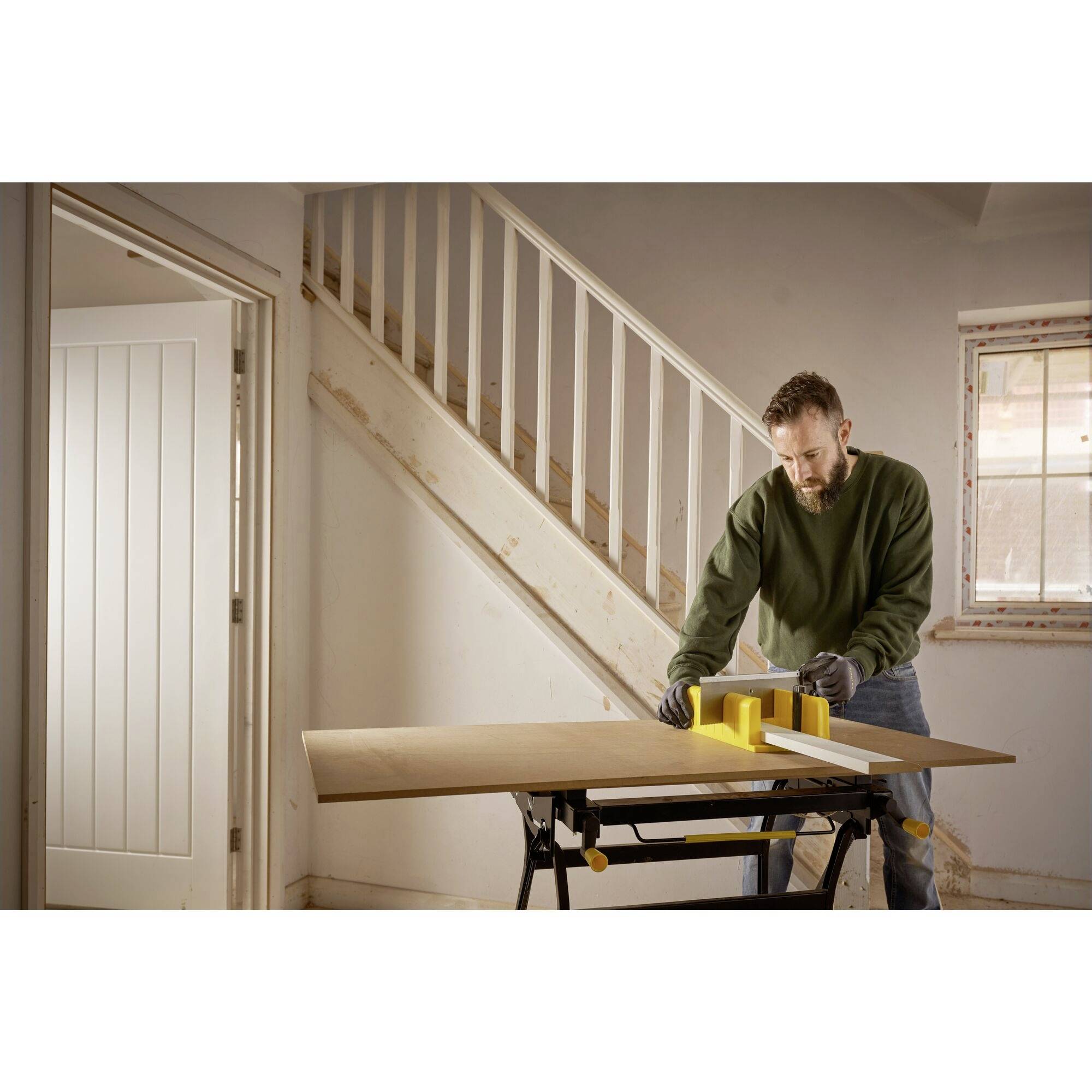 Ein Mann in einem leeren Raum schneidet ein Holzbrett mit einer Tischsäge. Im Hintergrund ist eine Treppe sichtbar.