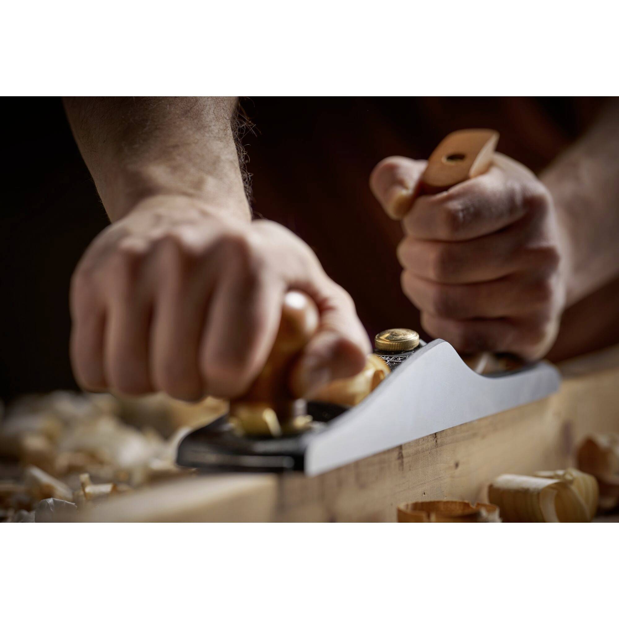 Eine Person hobelt ein Holzbrett mit einer Handhobelmaschine. Holzspäne liegen auf dem Tisch. Fokussiert auf die Arbeitsbewegung.