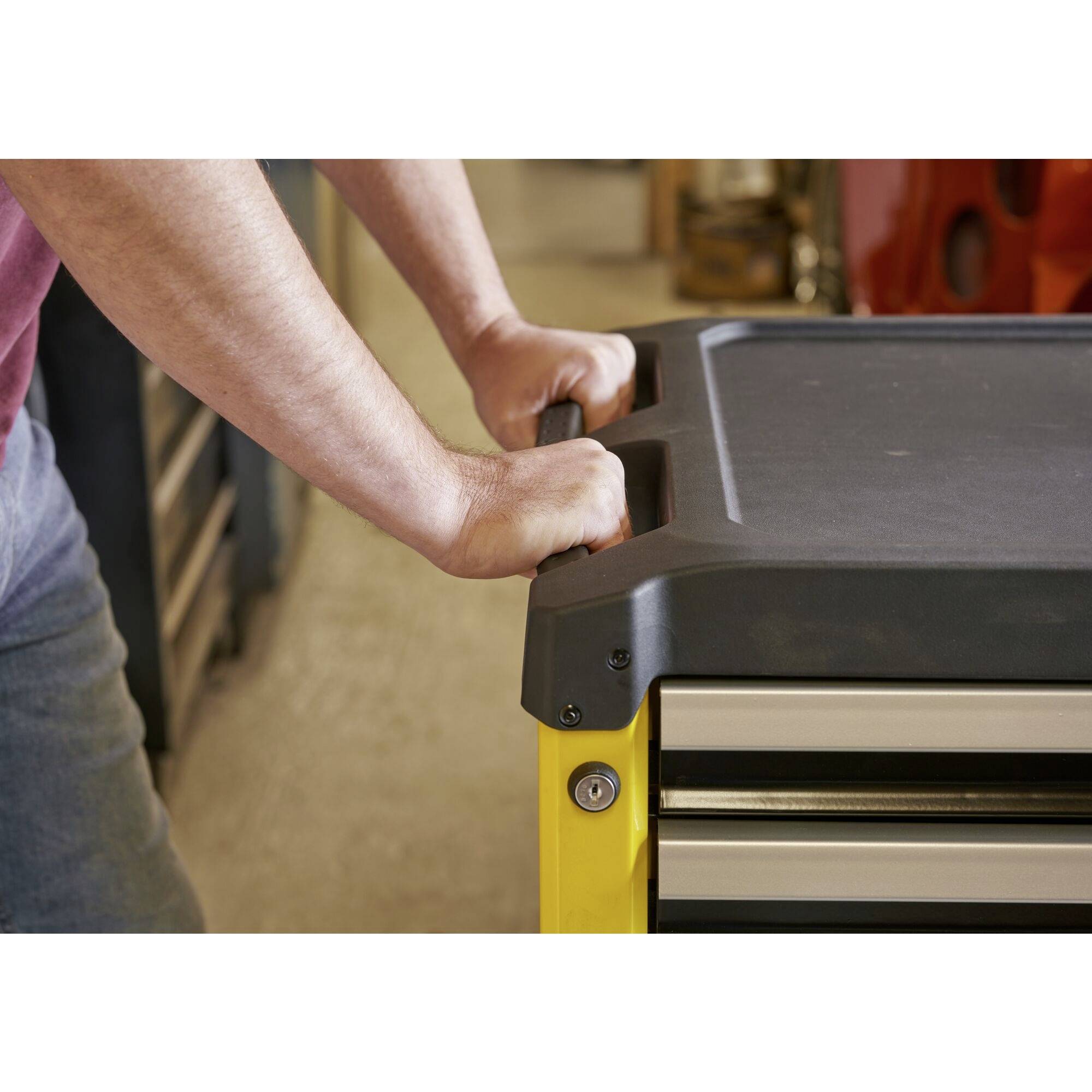 Eine Person stützt sich mit den Händen auf einen gelben Werkzeugwagen mit schwarzer Oberfläche. Im Hintergrund sind Werkzeuge zu sehen.