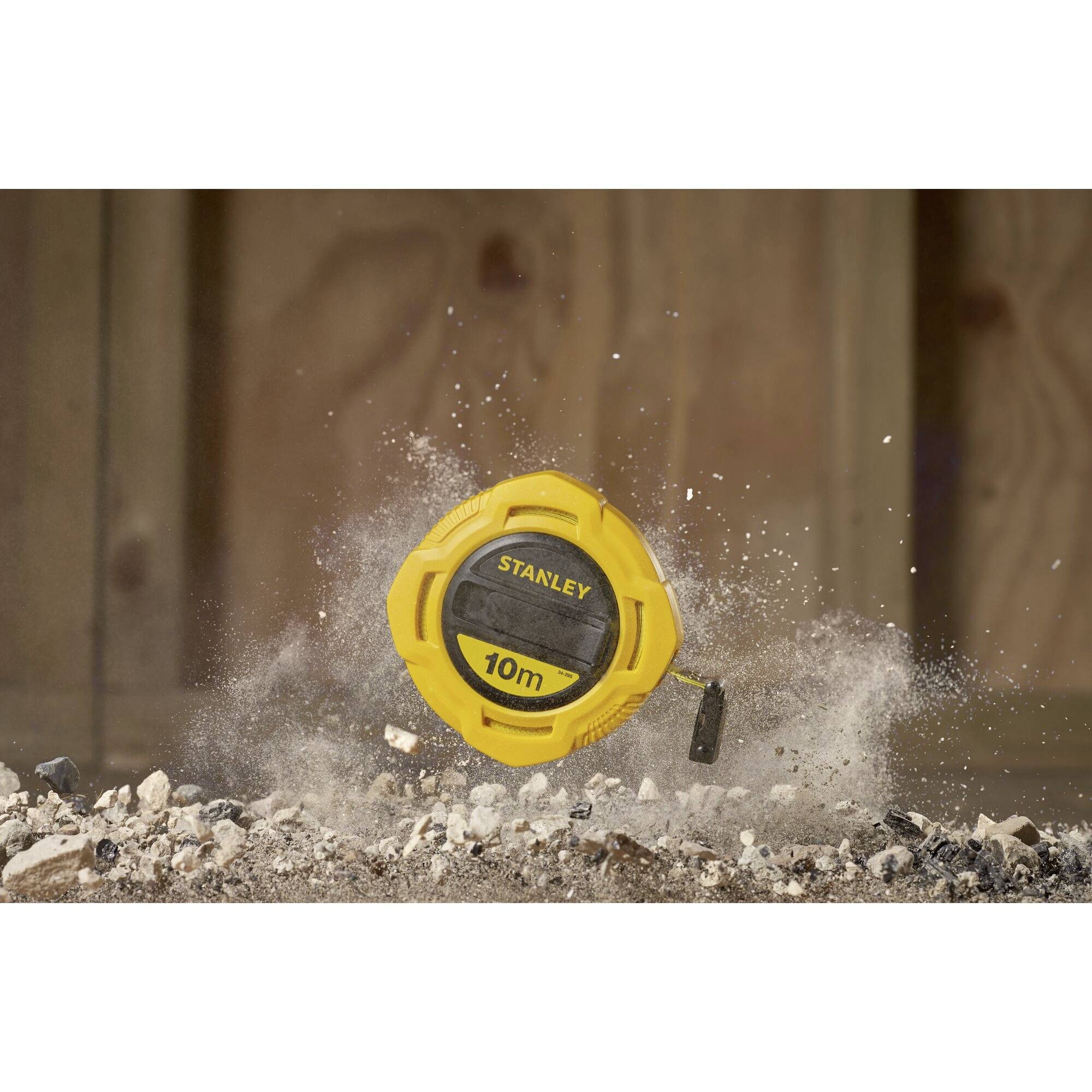 Ein gelbes Maßband 'Stanley' mit 10m Markierung liegt auf staubigem Boden, umgeben von aufgewirbeltem Staub und Trümmern.