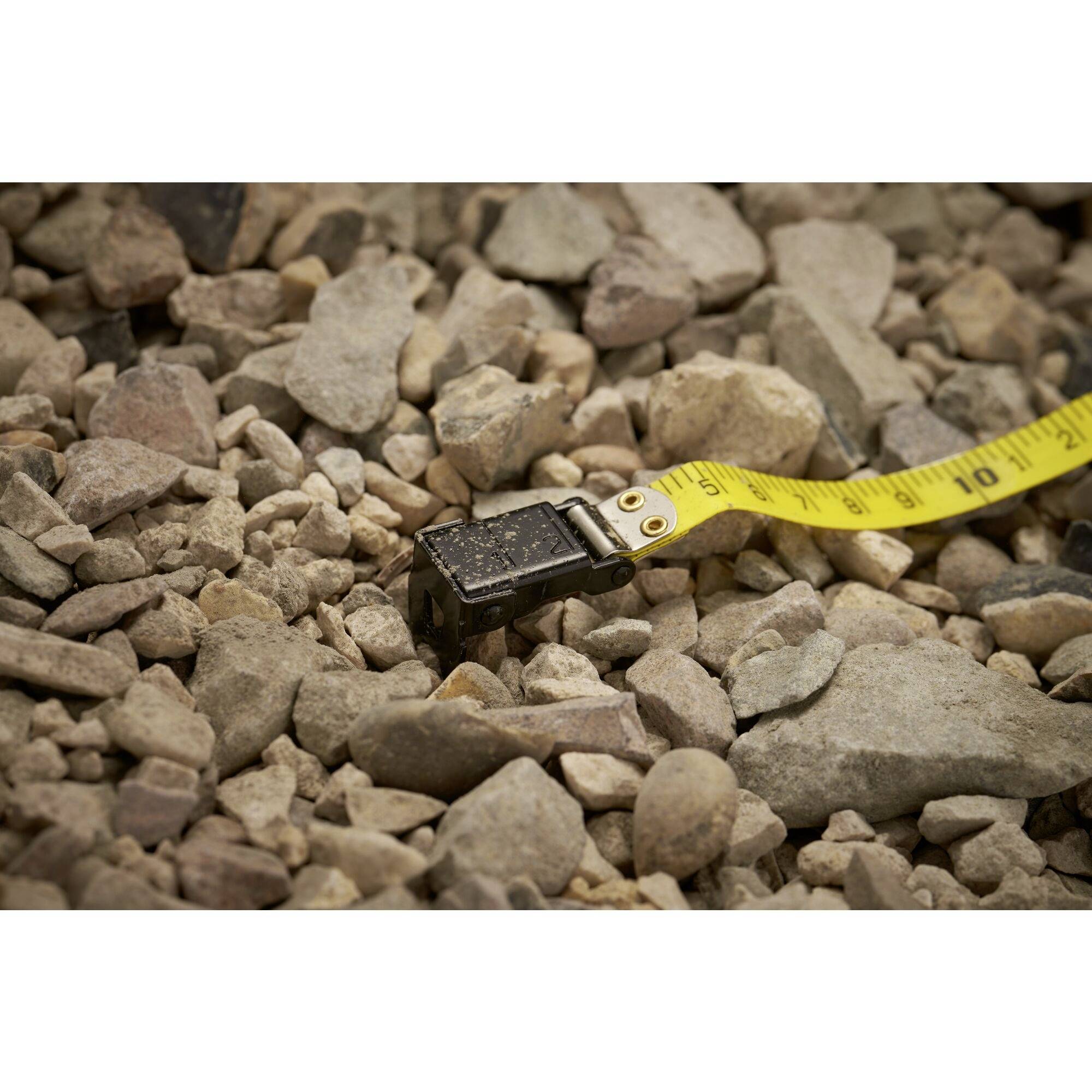 Ein Maßband liegt auf einem Boden aus grobem Kies. Das Maßband zeigt eine Länge, die auf den Steinen ruht.