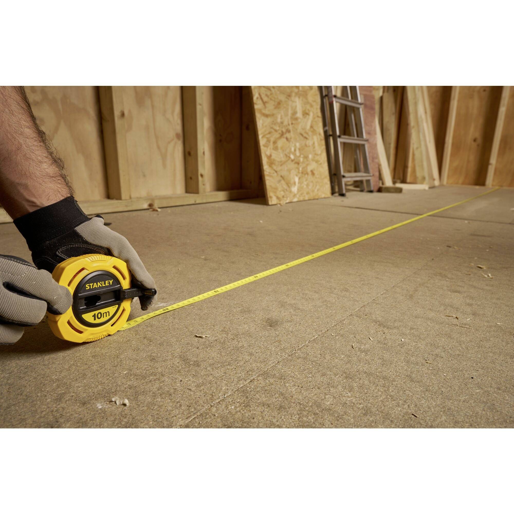 Eine Person misst mit einem gelben Maßband eine Distanz auf einem Boden in einem teilweise erbauten Holzraum.